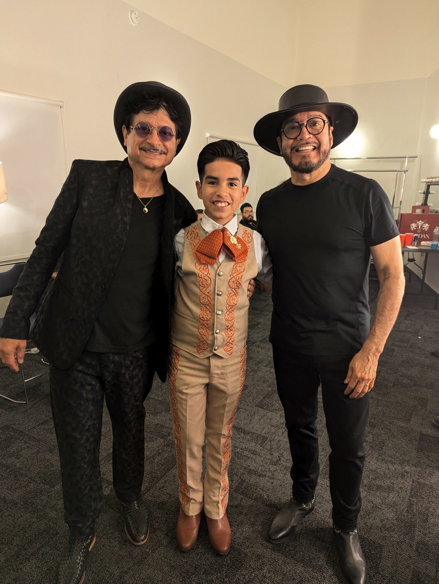 Three people standing indoors, two men and a boy, smiling, dressed in stylish clothing and hats.