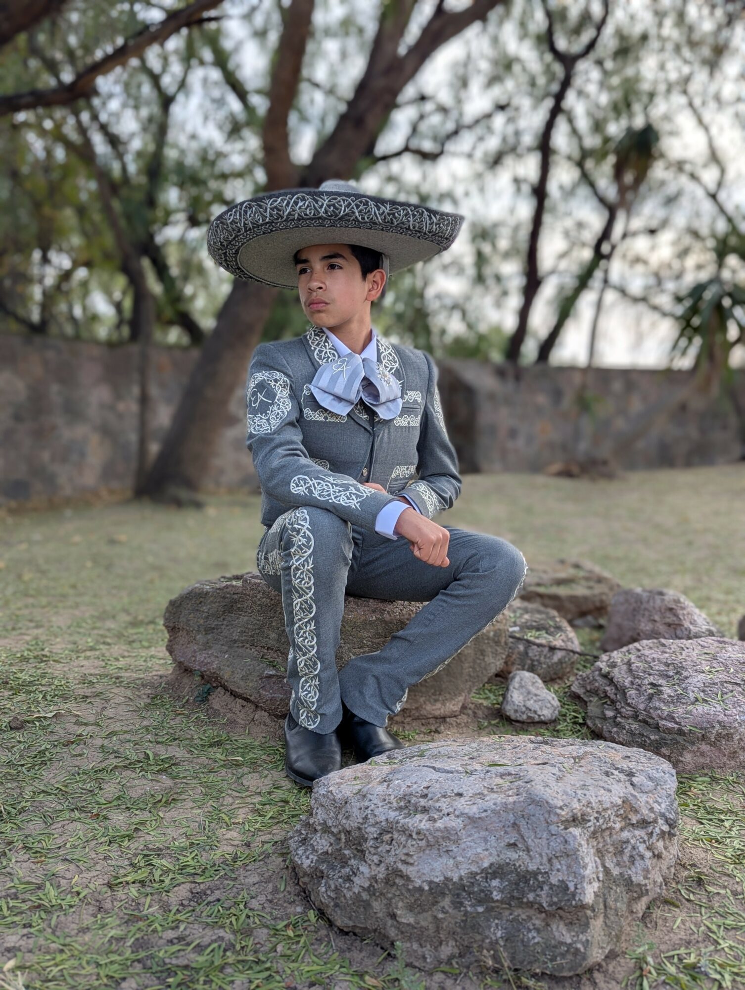 Child dressed as a mariachi, sitting on rocks outdoors, wearing a large sombrero and traditional outfit, with trees in background.