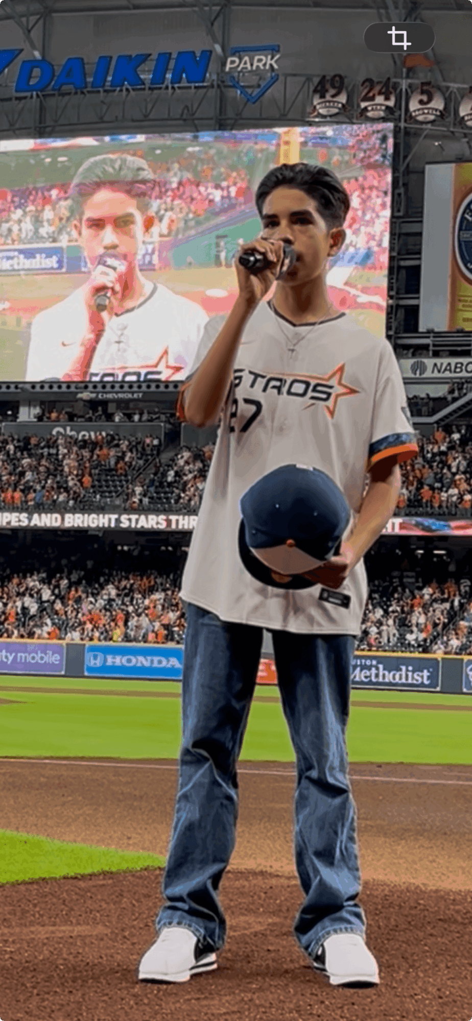 Young man holding a baseball and microphone on a baseball field, stadium in background, wearing a white jersey and jeans.
