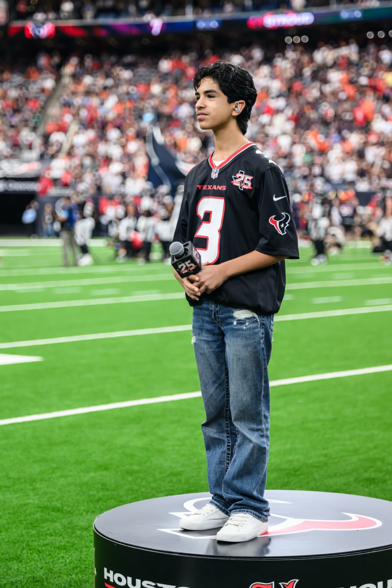 Young man standing on a circular platform on a football field, wearing a black jersey and jeans, with a crowd in the background.
