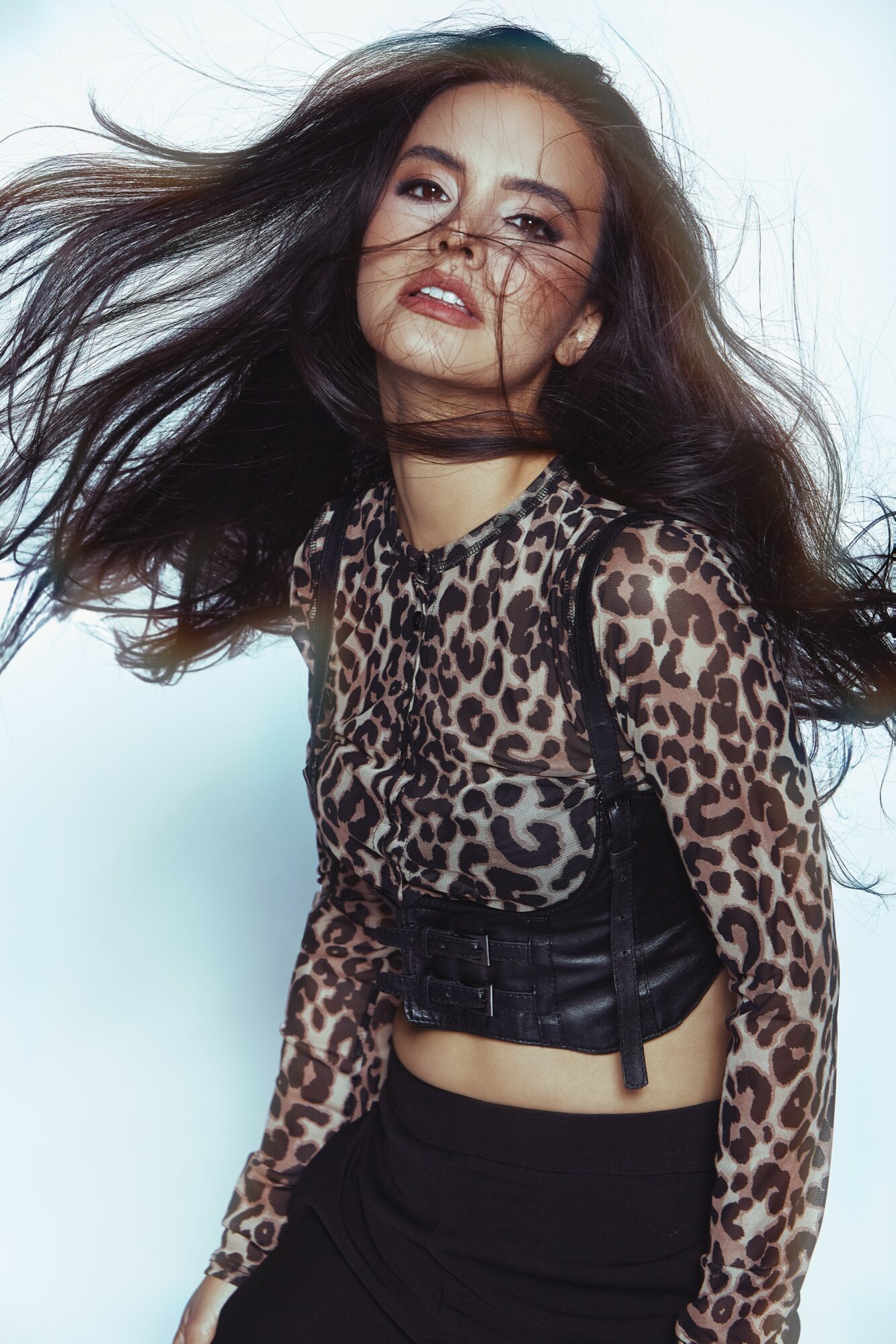 Young woman with long hair wearing a leopard print top and black pants, looking at the camera with wind-blown hair.