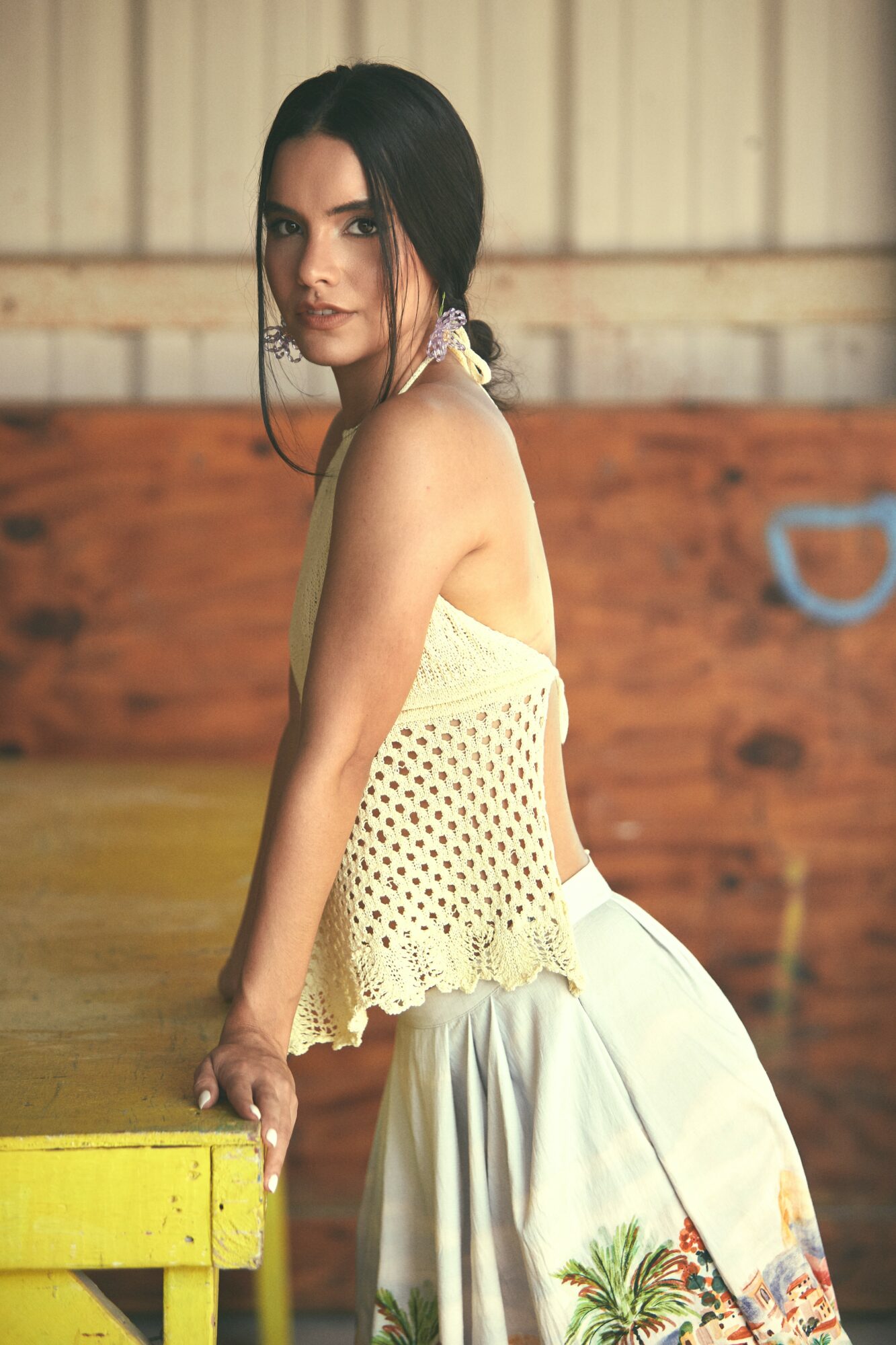 Young woman with dark hair in a ponytail, wearing a sleeveless crochet top and white skirt with palm tree print, leaning on a yellow table indoors.