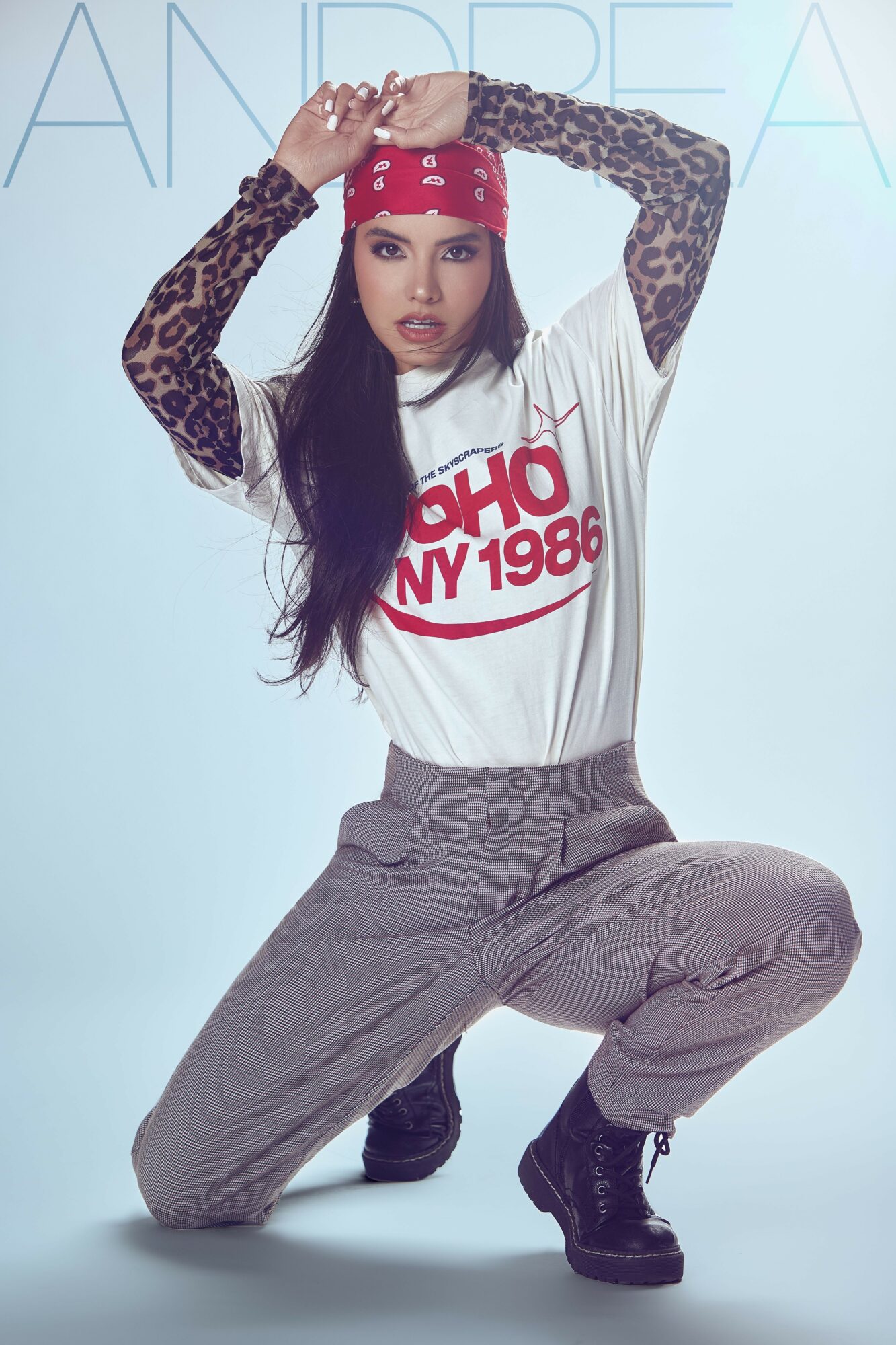 Woman with long dark hair wearing a red bandana, white graphic t-shirt, leopard print sleeves, gray pants, and black boots, squatting.