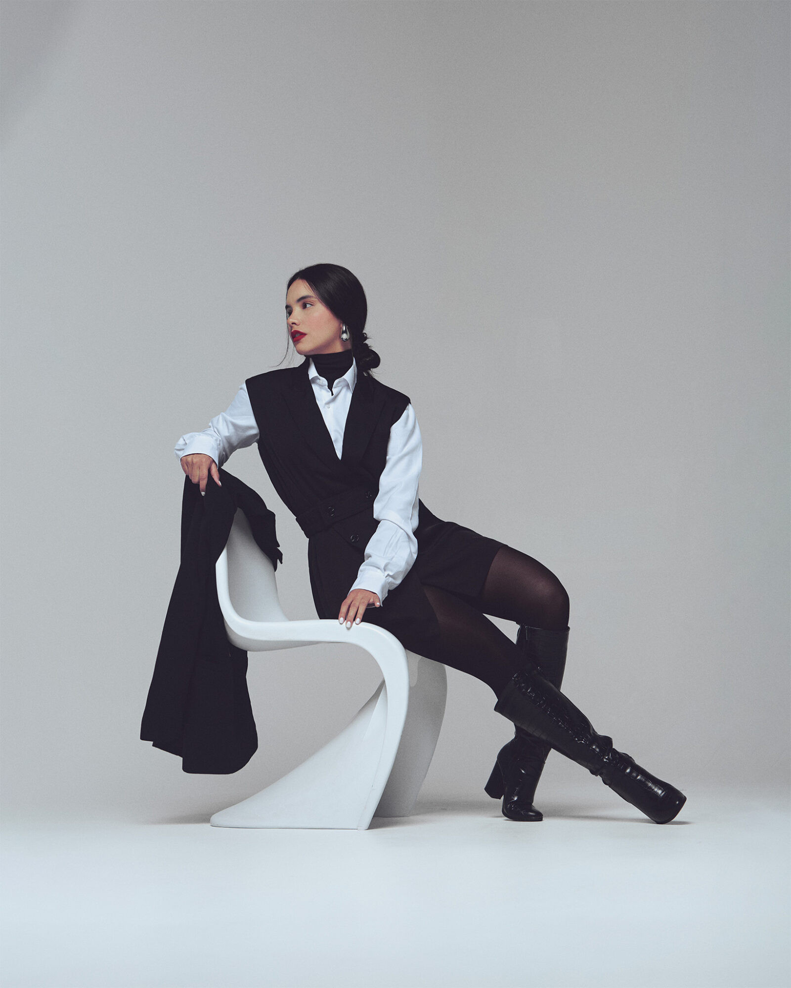 Woman sitting on a modern white chair, wearing a black vest, white shirt, black tights, and boots, against a plain background.