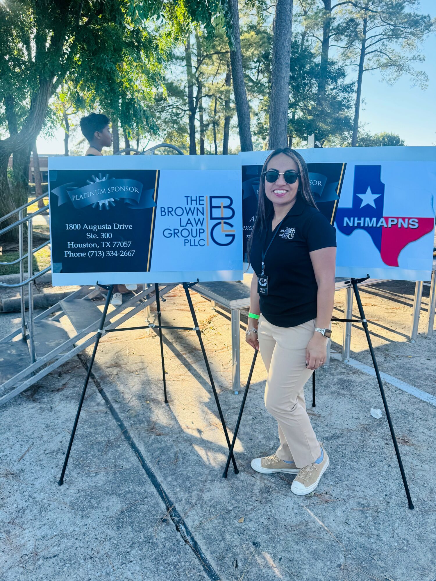 Woman standing outdoors in front of a large display board, smiling, wearing sunglasses, black shirt, and beige pants.