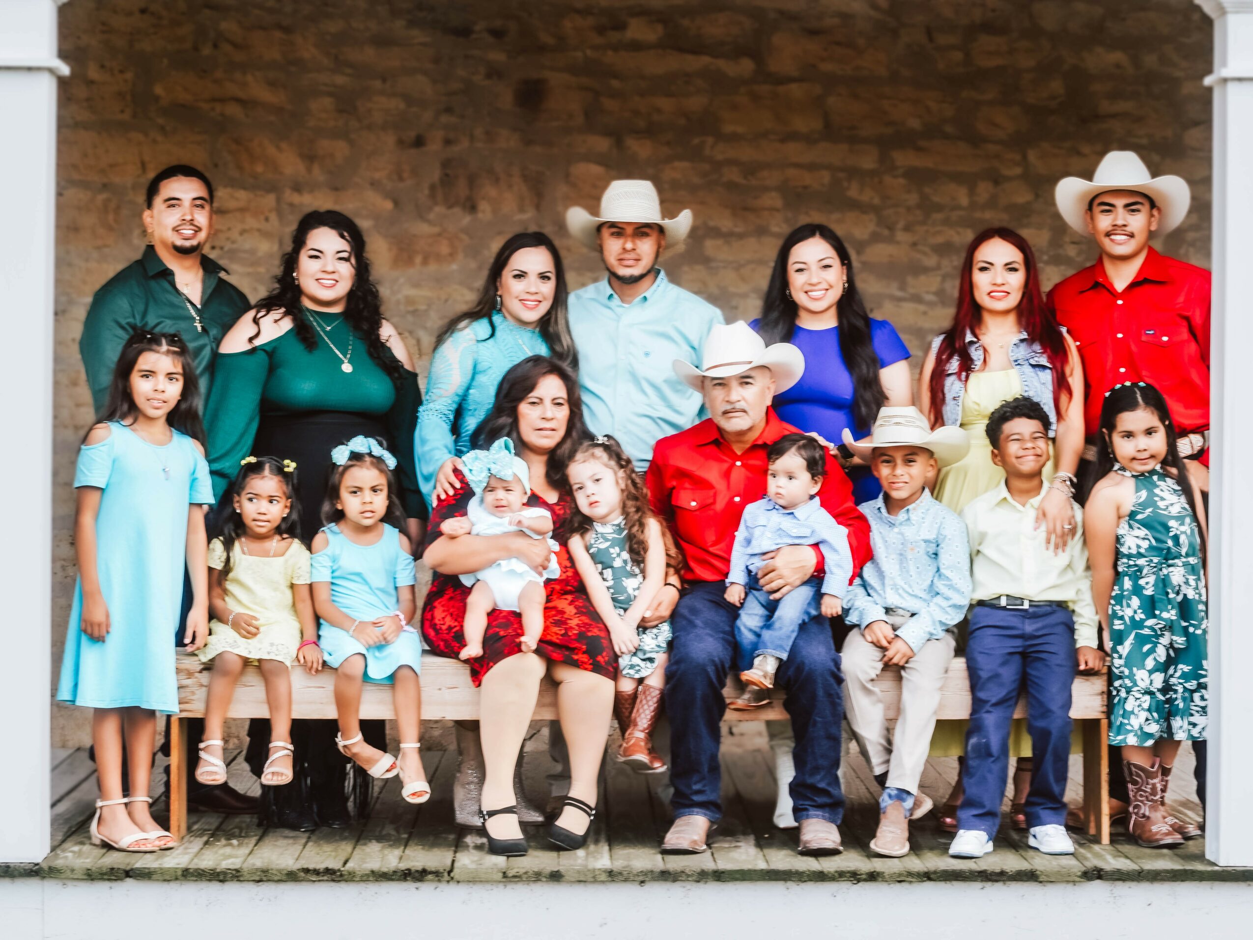Group of children and adults posing outdoors against a brick wall, some wearing cowboy hats and colorful clothing.