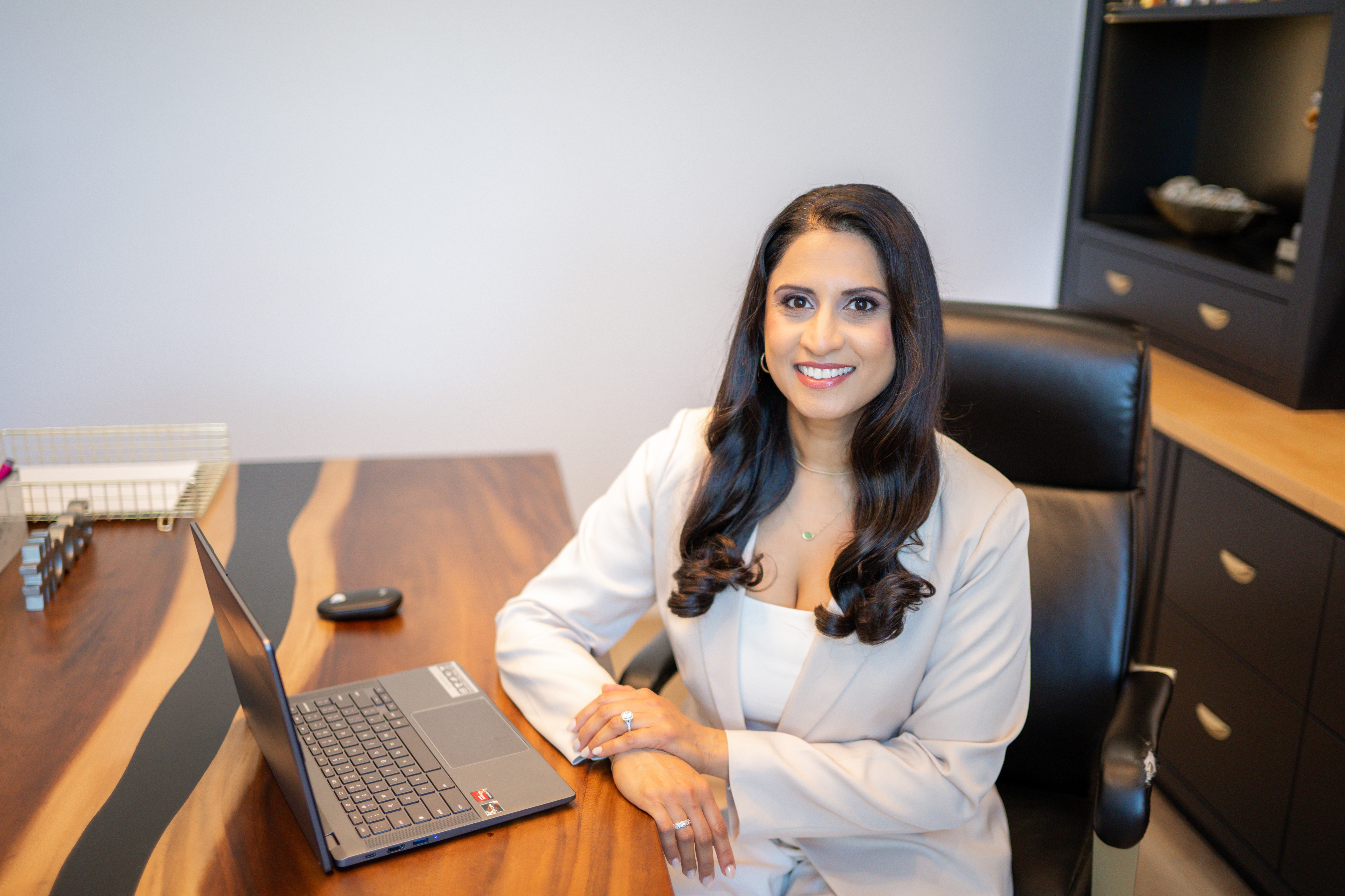 Woman with long dark hair in a white blazer sitting at a desk with a laptop, smiling, in an office setting.