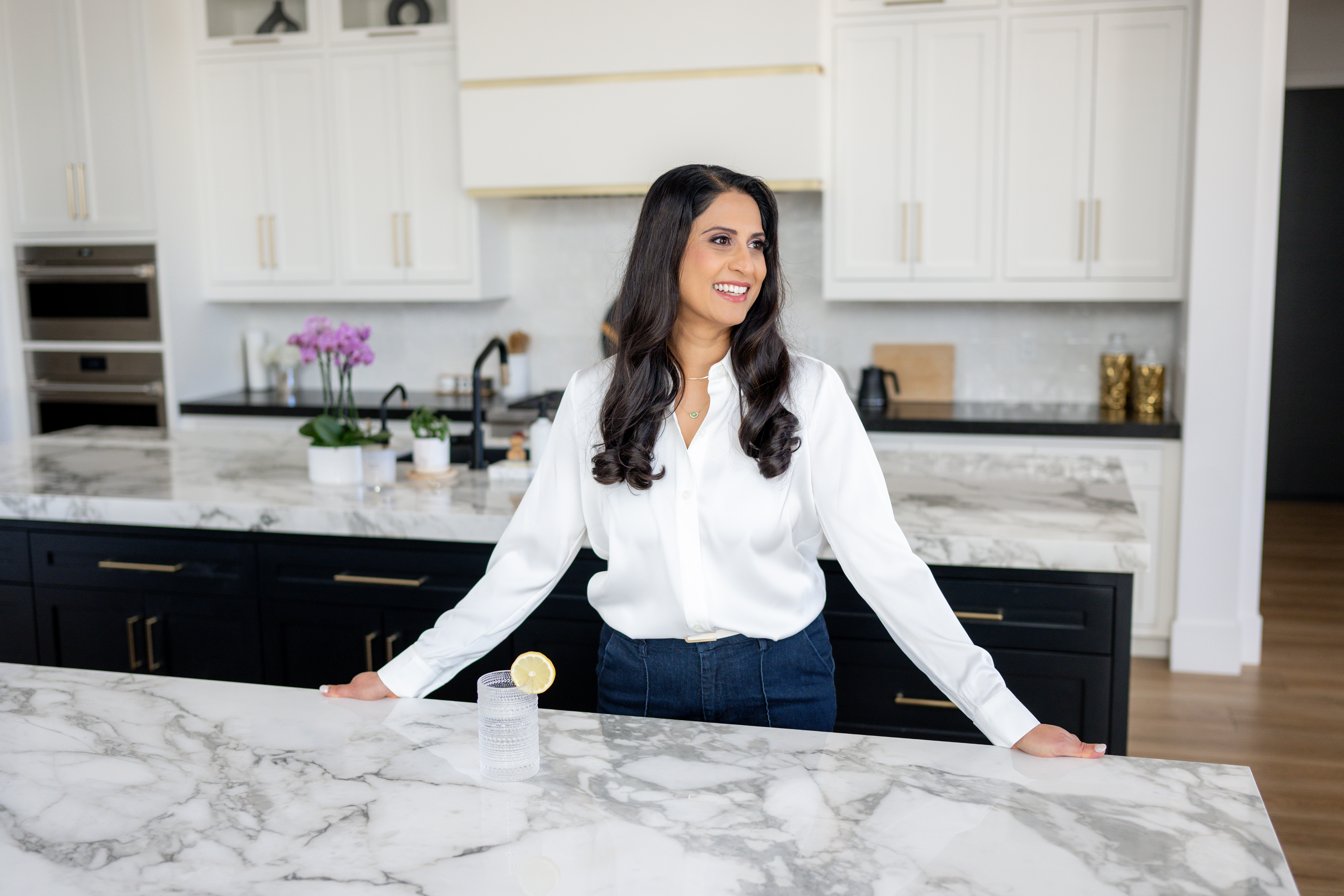 Woman with long dark hair smiling, standing behind a marble kitchen counter, wearing a white blouse and jeans, in a modern kitchen.