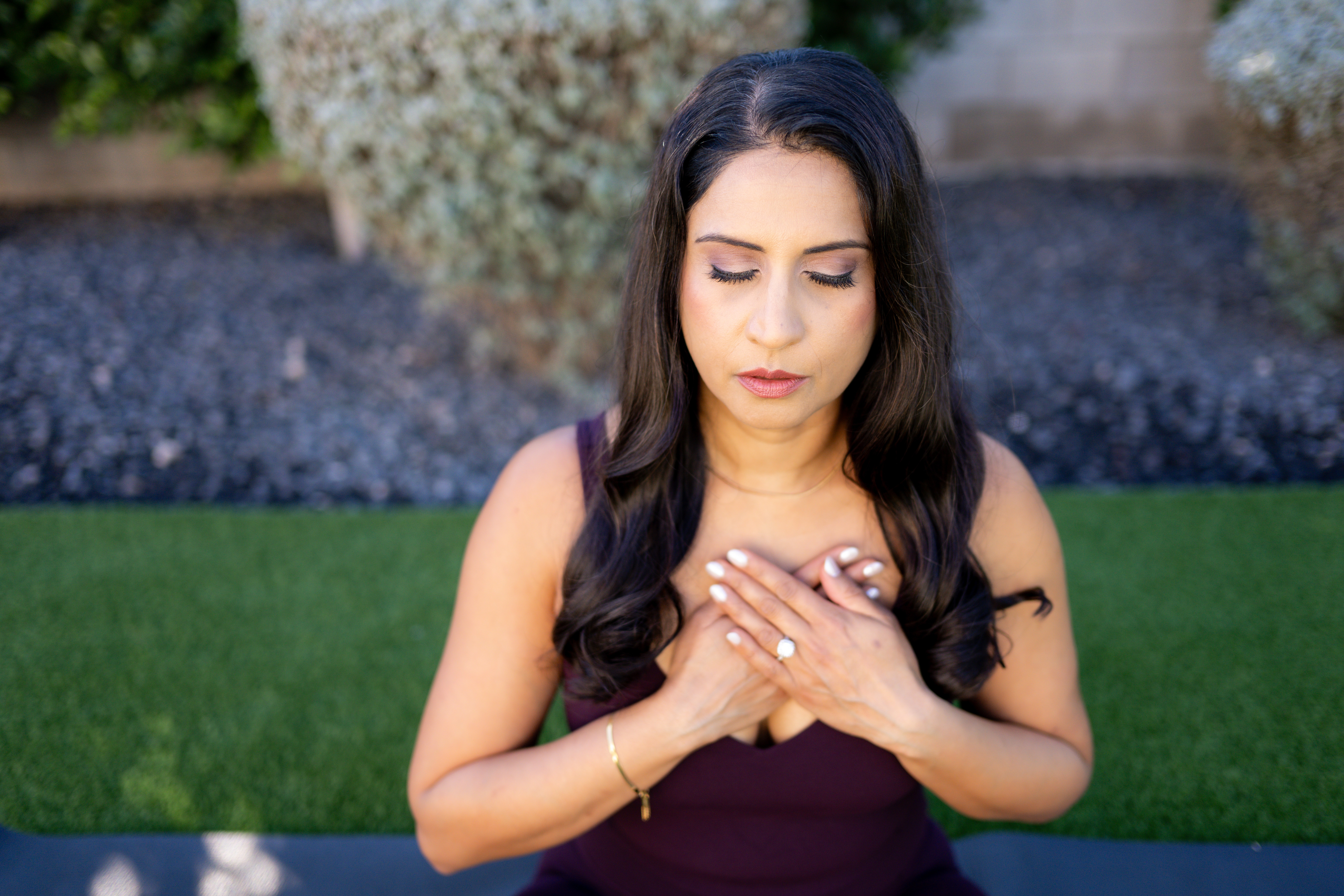 Young woman with long dark hair, wearing a sleeveless dark top, holding her hands over her chest outdoors.