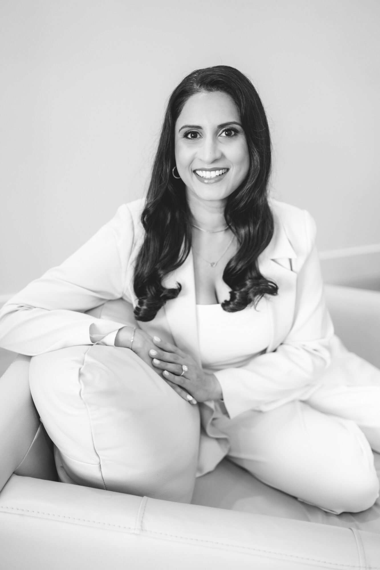 Woman with long dark hair smiling, sitting on a couch, wearing a light-colored outfit, in a plain room.