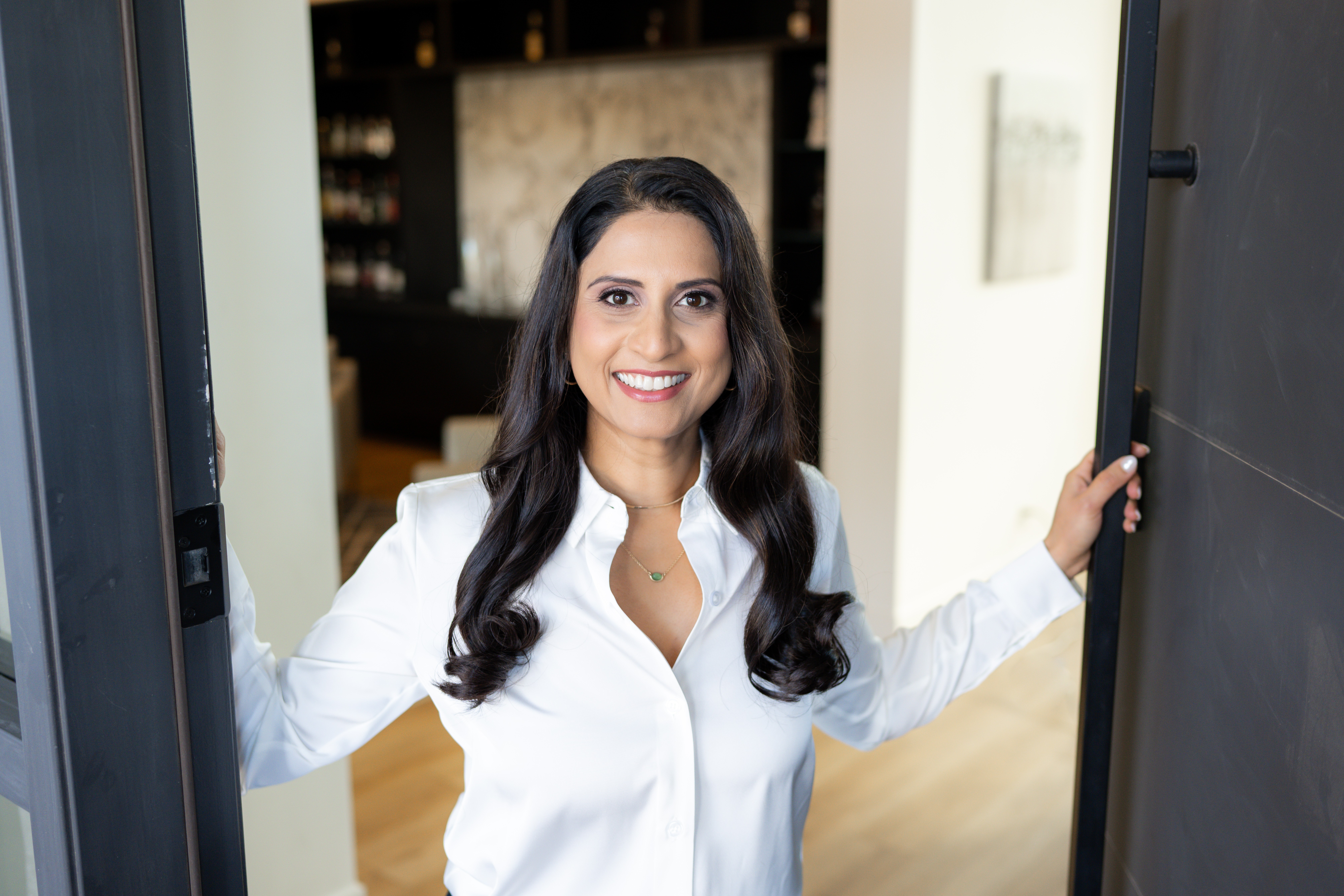 Woman with long dark hair smiling, wearing a white shirt, standing in doorway indoors.