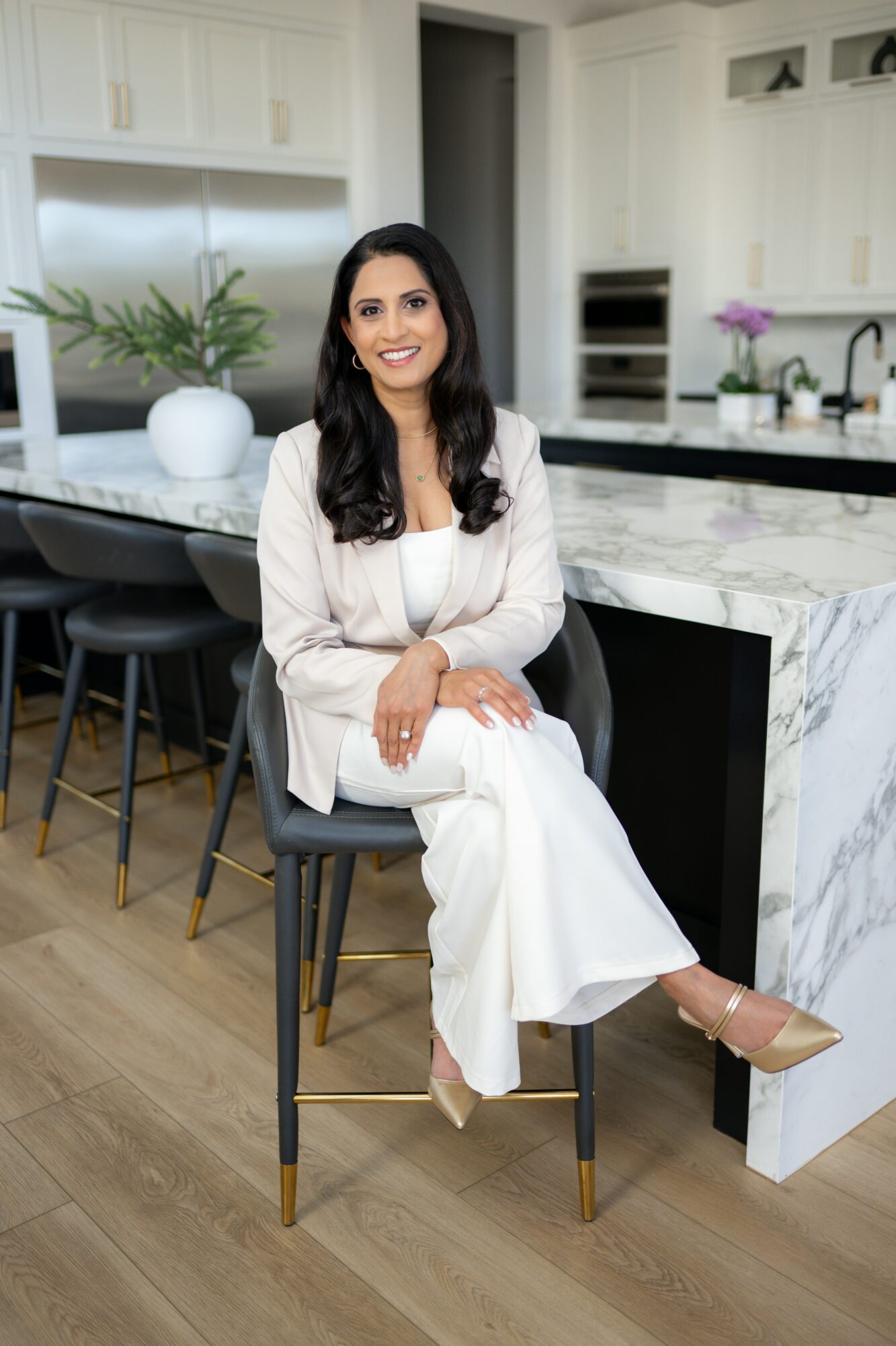 Woman sitting on a barstool in a modern kitchen with white cabinets, marble countertops, and a potted plant.