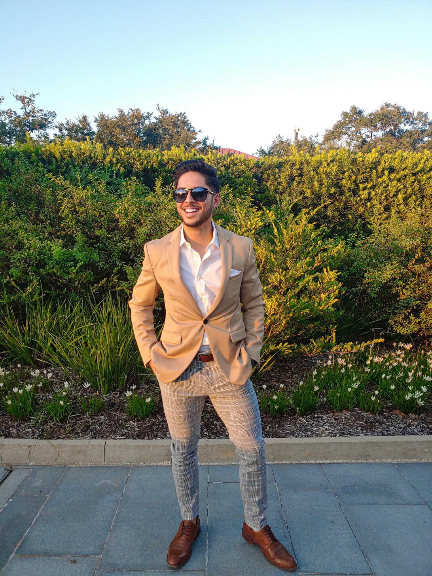 Man in beige blazer, white shirt, patterned pants, and sunglasses standing outdoors with greenery behind him.