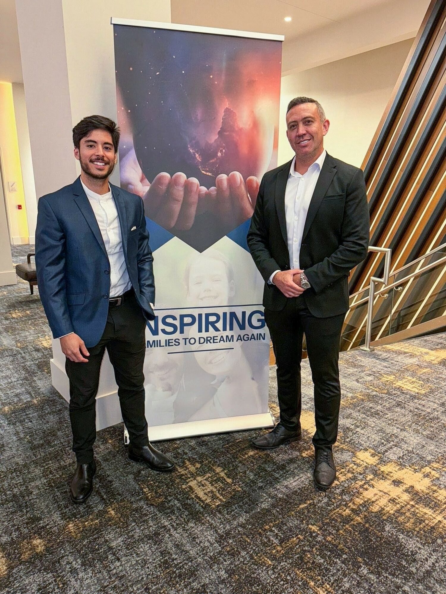 Two men in suits standing next to a banner with a galaxy and hand image, smiling indoors.