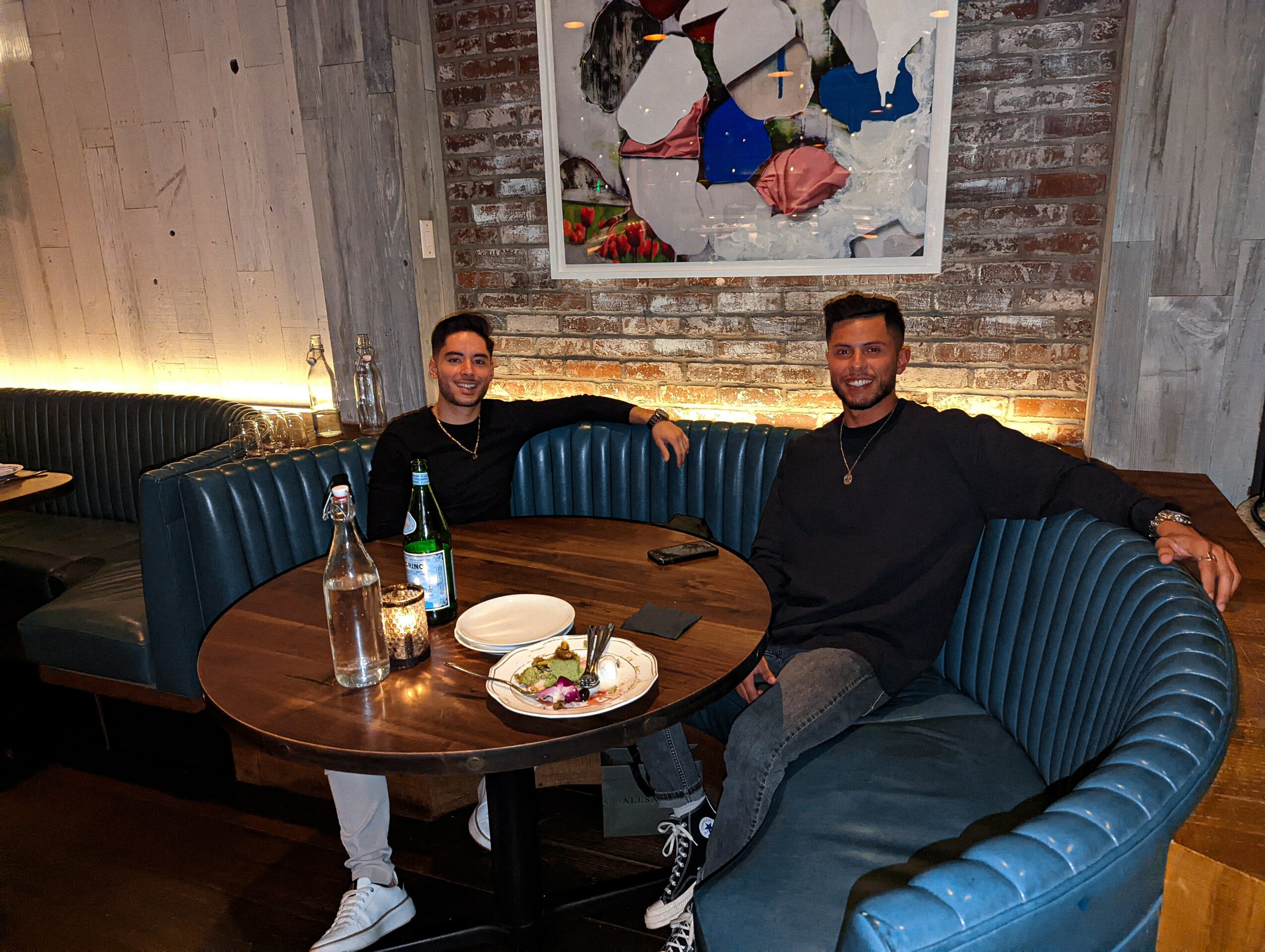 Two men sit at a round table in a restaurant, smiling, with food and drinks on the table, brick wall background.