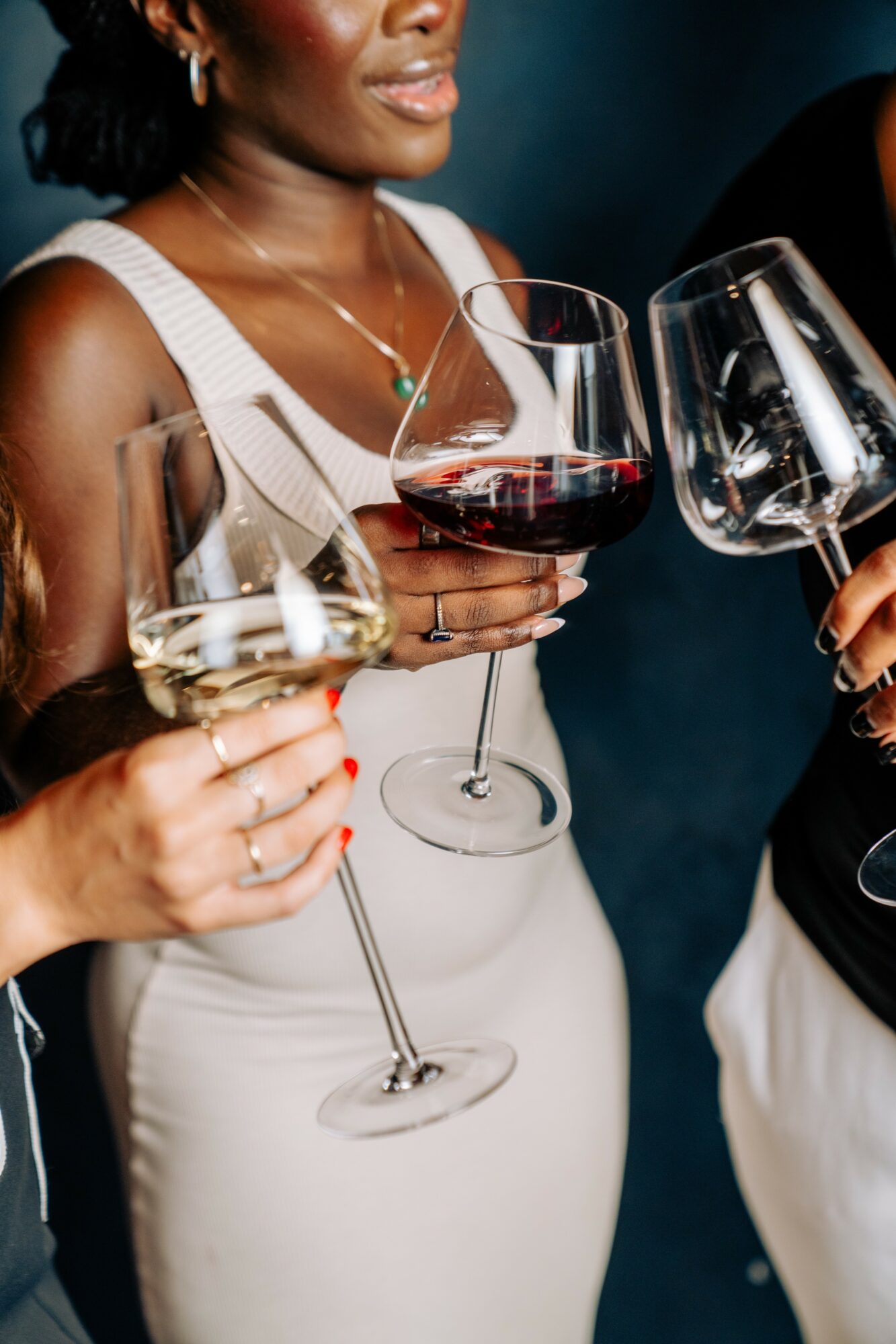 Three women holding wine glasses, one with red wine, at a social gathering, close-up of upper bodies and hands.