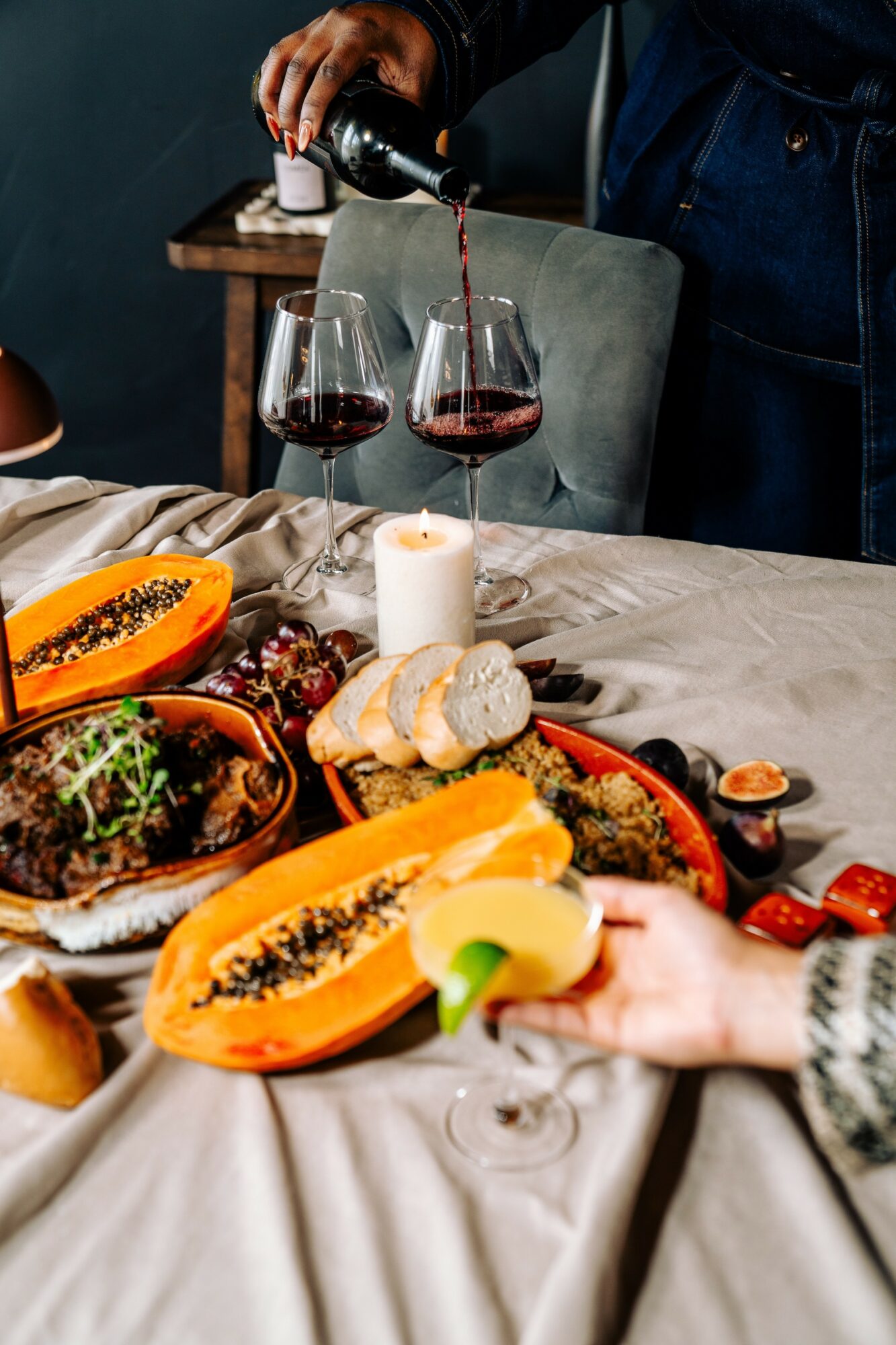 Person pouring red wine into glasses on a table with fruit, bread, and a candle, with a hand reaching for fruit.