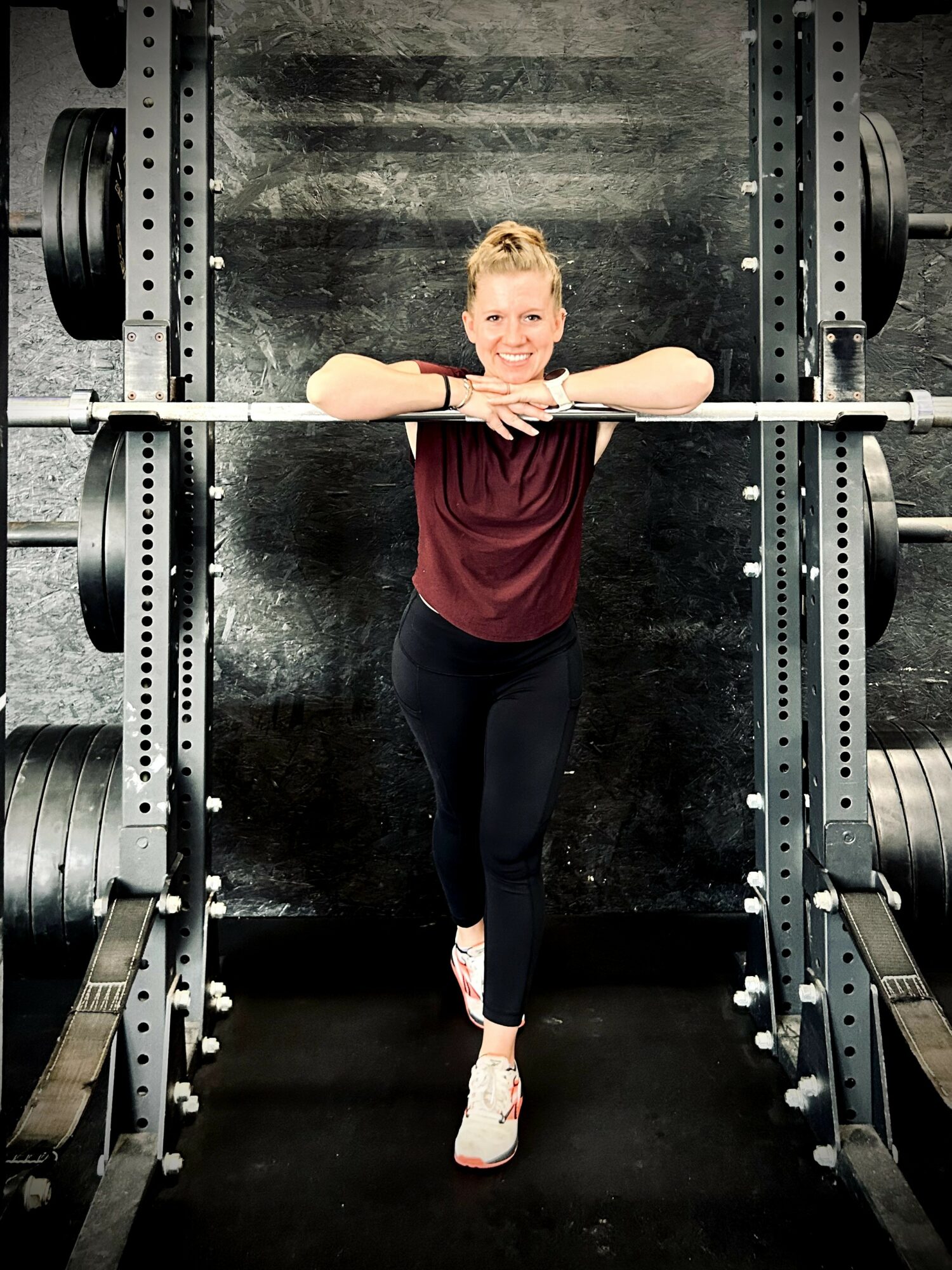 Young woman standing in a gym, leaning on a barbell, smiling, with weightlifting equipment around her.