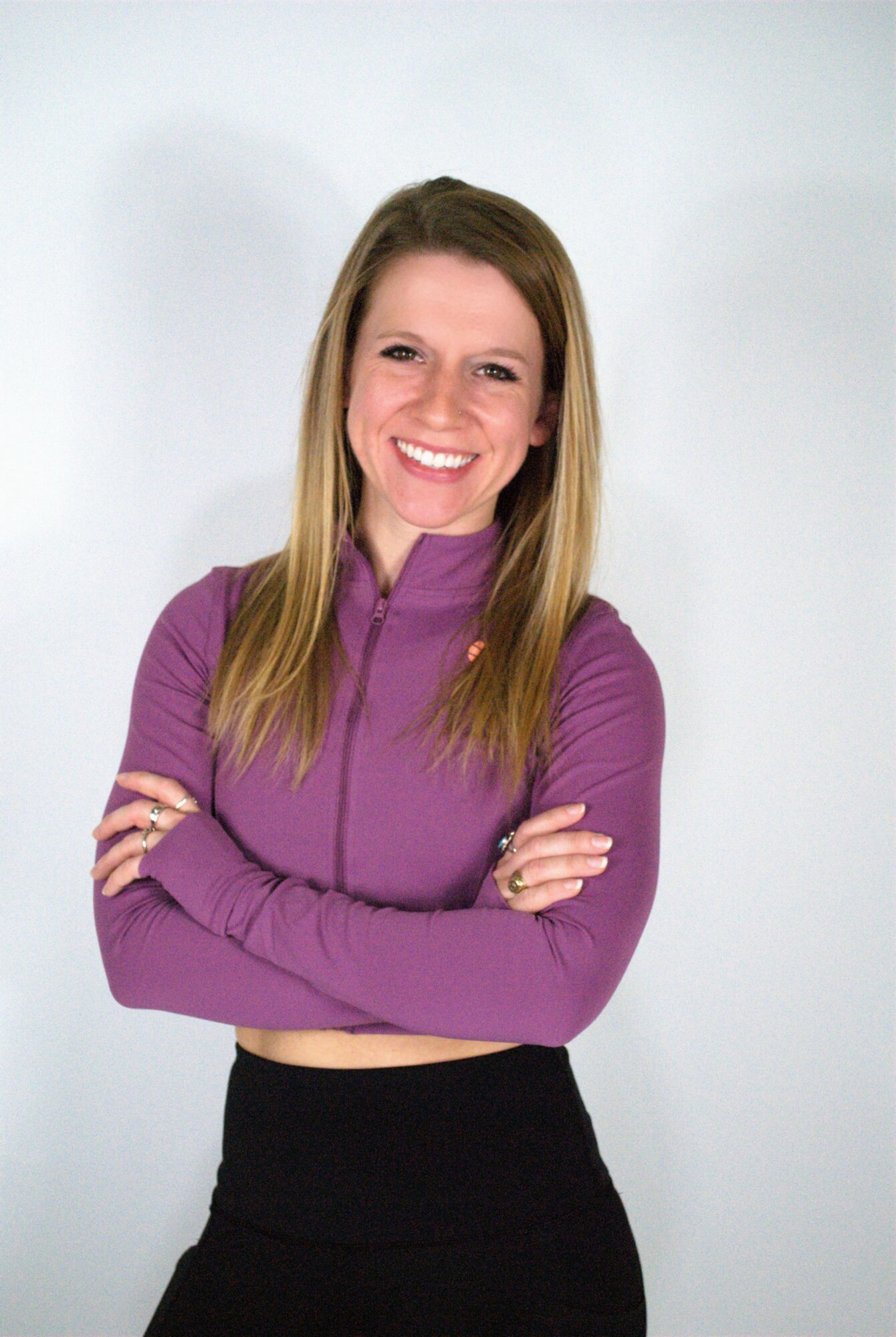 Young woman with long hair smiling, arms crossed, wearing a purple top and black skirt, standing against a light background.