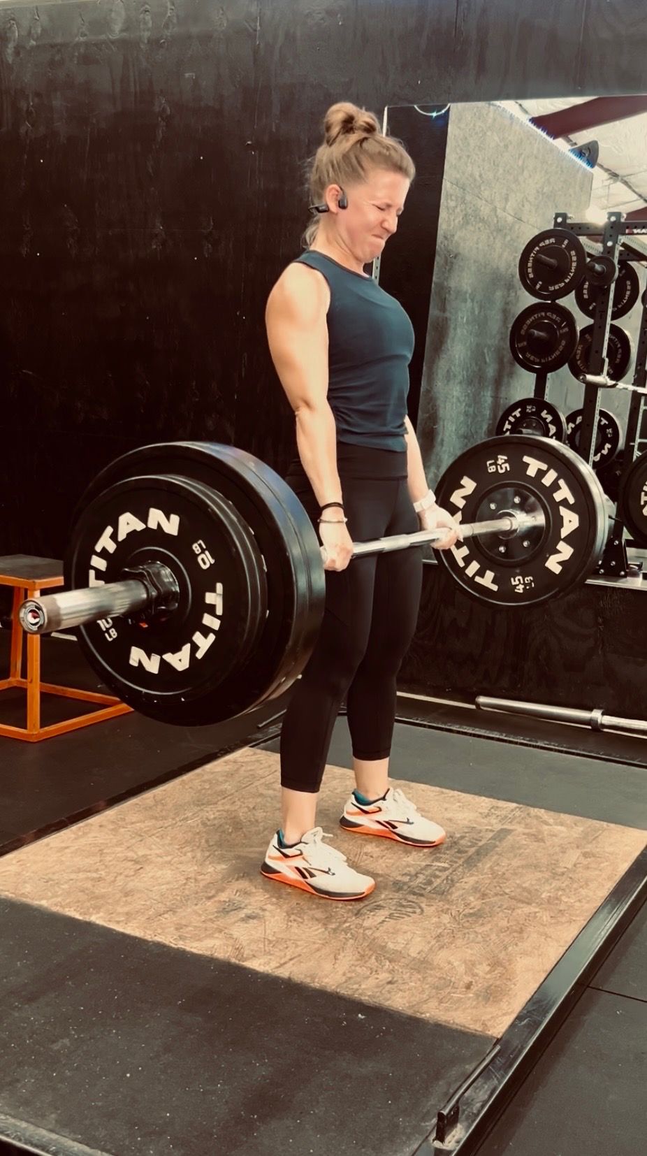 Woman lifting a barbell in a gym, standing on a mat, with weight plates and equipment in background.