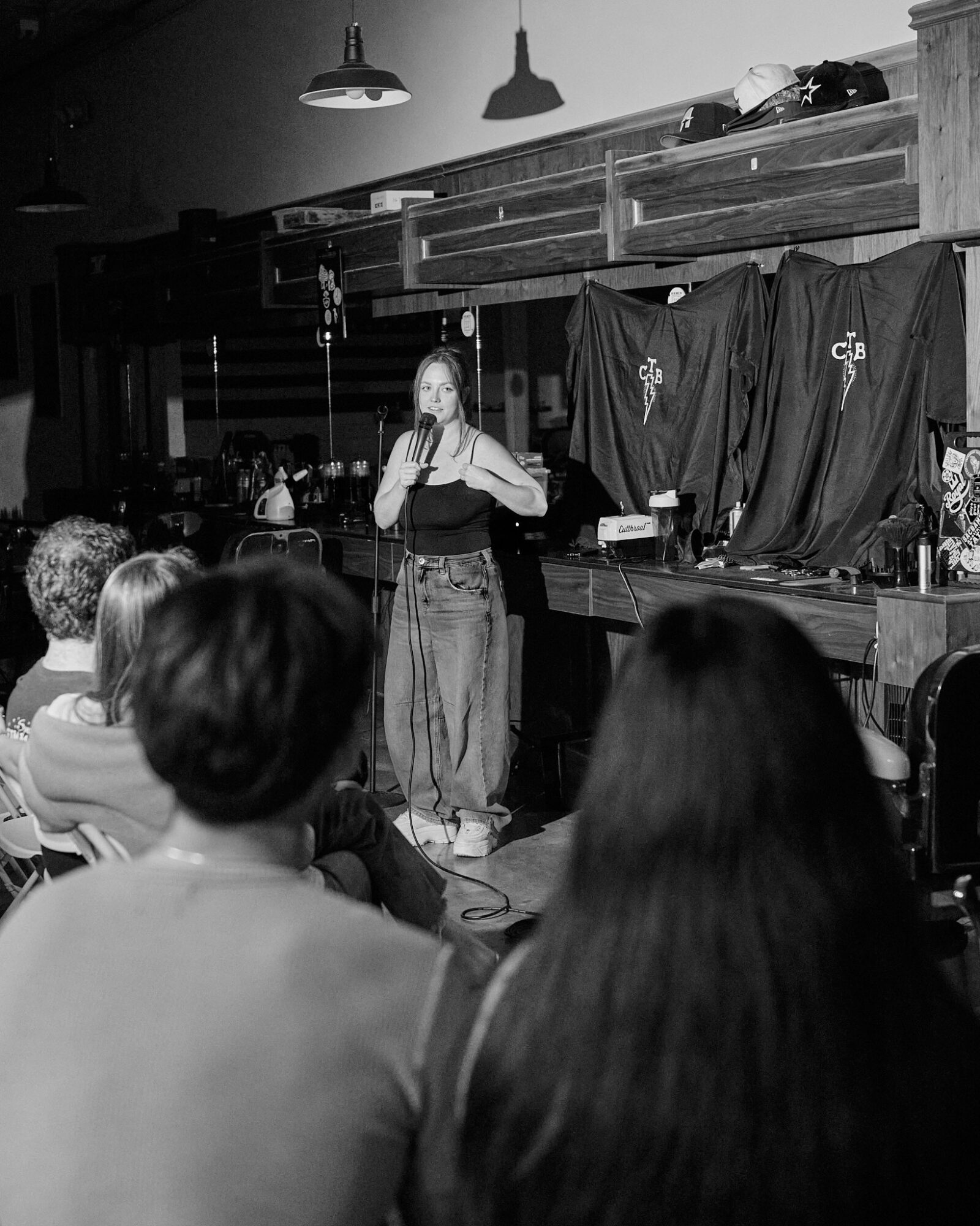 Person standing on stage speaking to an audience in a dimly lit room with hanging lights and black curtains behind.
