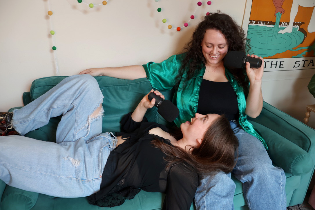 Two women are relaxing on a green couch, one lying down and the other sitting, smiling and talking, in a decorated room.
