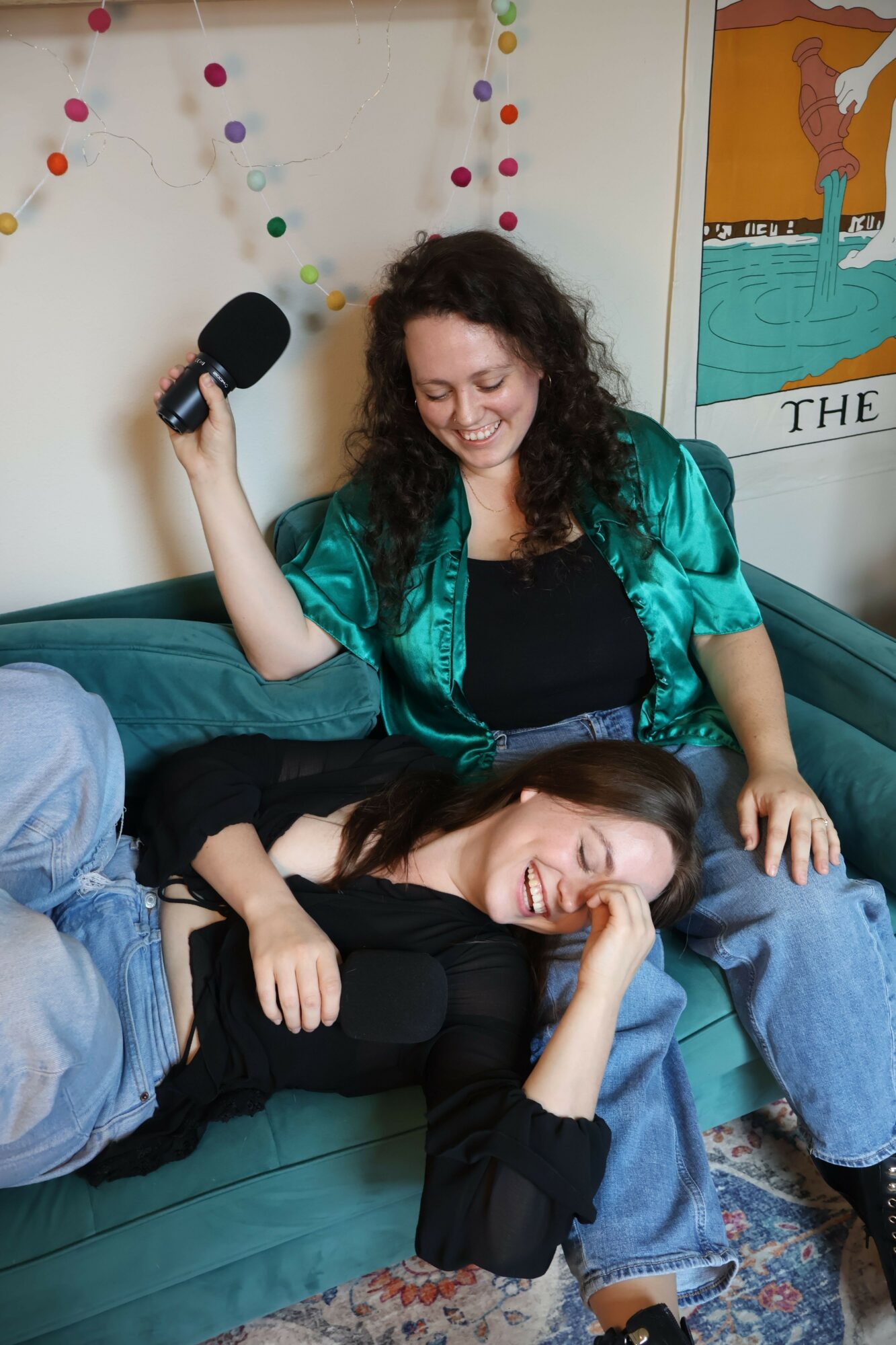 Two women sitting on a couch, one holding a microphone, smiling, with colorful decorations in the background.