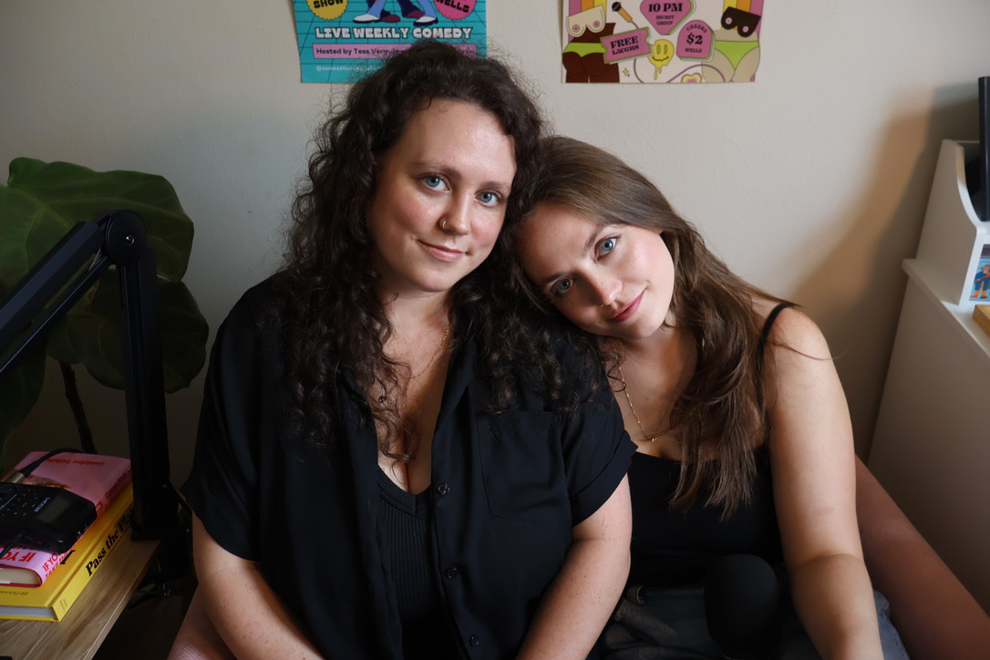 Two women sitting close together indoors, one with curly hair and the other with straight hair, smiling at the camera.