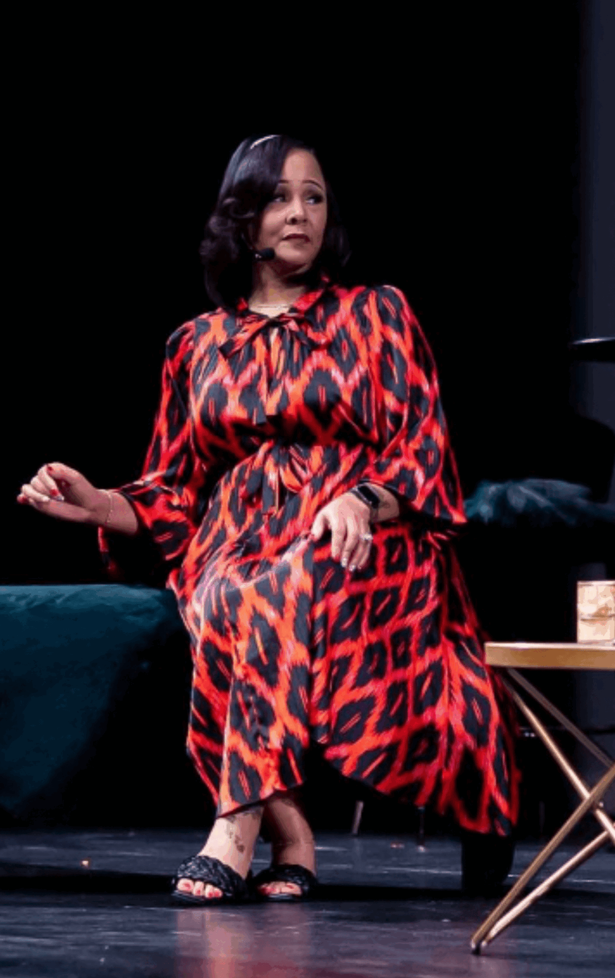 Woman sitting on a stage, wearing a red and black patterned dress, with a dark background and a small table nearby.
