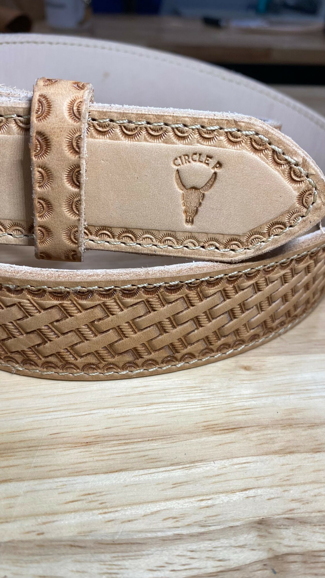 Close-up of a woven belt with a beige leather tip, embossed with a logo and decorative stitching, on a wooden surface.