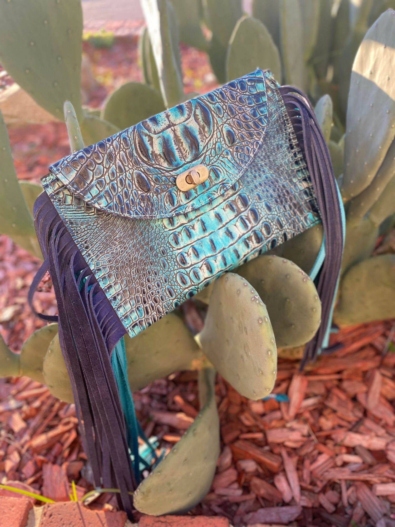 Colorful crocodile-patterned wallet with purple sides, resting on rocks among cactus plants in a garden.