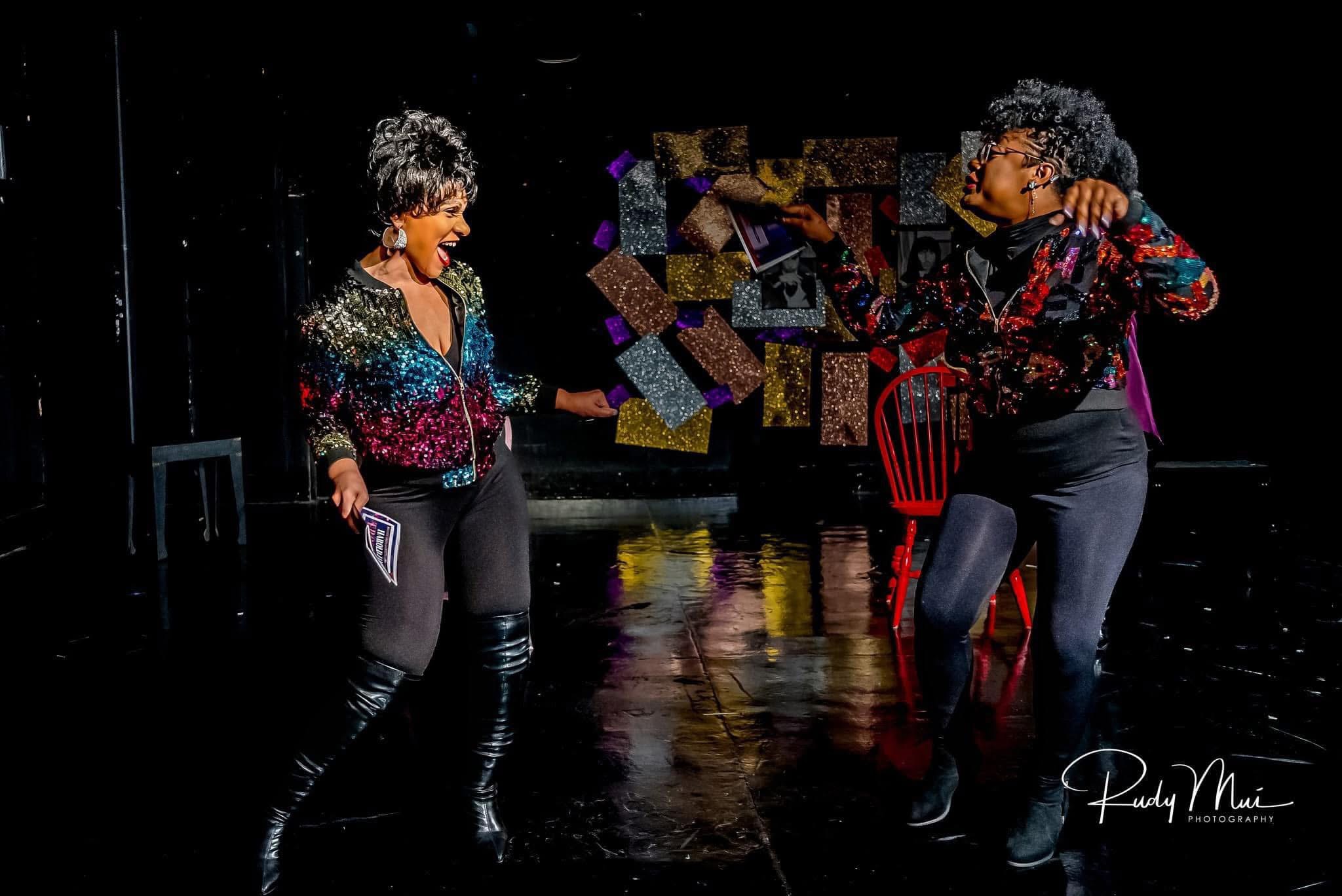 Two women dance on a stage with a dark background and colorful decorations, wearing patterned tops and black pants.
