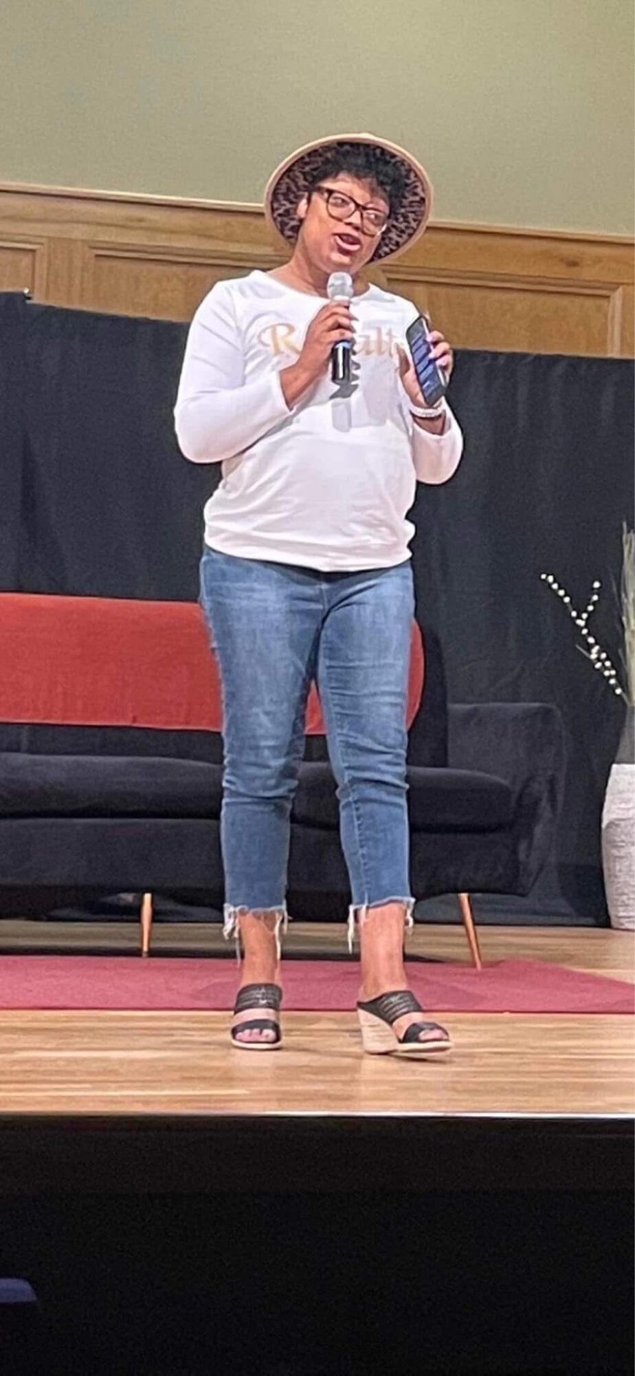 Person standing on stage holding a microphone and a phone, wearing glasses, a hat, white sweater, and jeans.