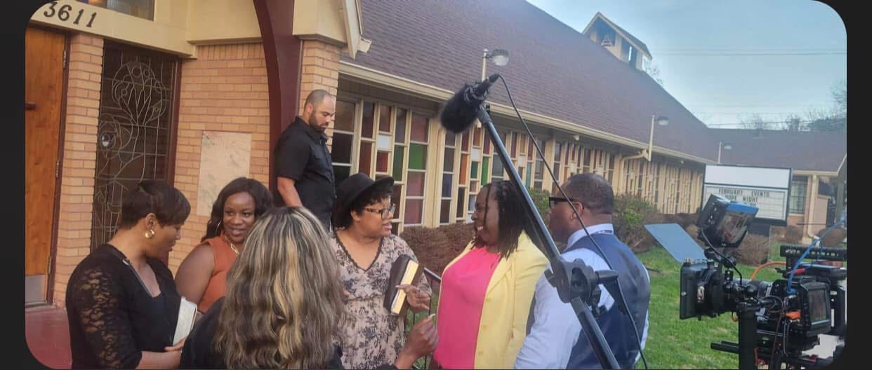Group of people gathered outdoors near a building, with a camera and boom microphone visible, possibly filming or interviewing.