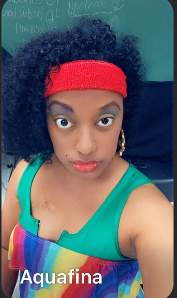 Woman with curly hair wearing a red headband and green top, looking at camera, with a rainbow-colored strap and earrings.