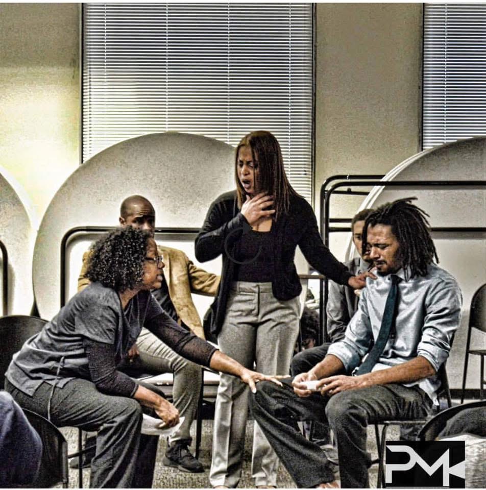 Group of five people in a room, one woman standing and speaking, others seated and listening, with large windows behind.