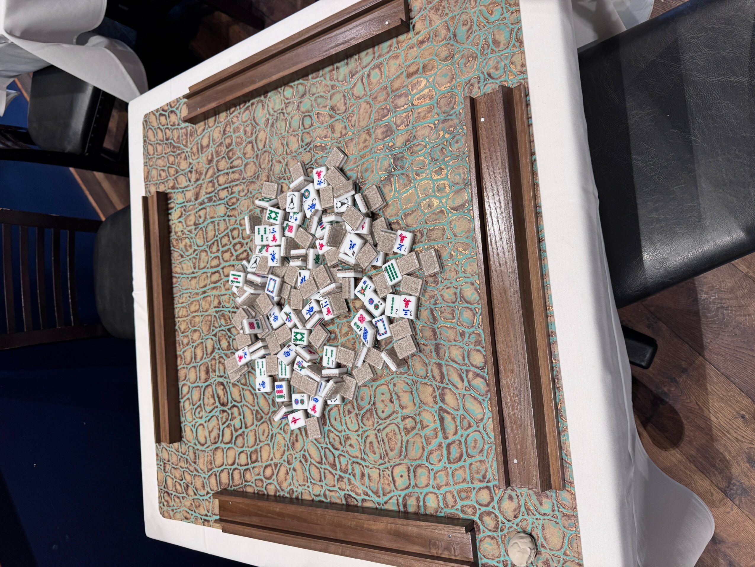 Mahjong tiles scattered on a patterned table with a wooden border, viewed from the side.