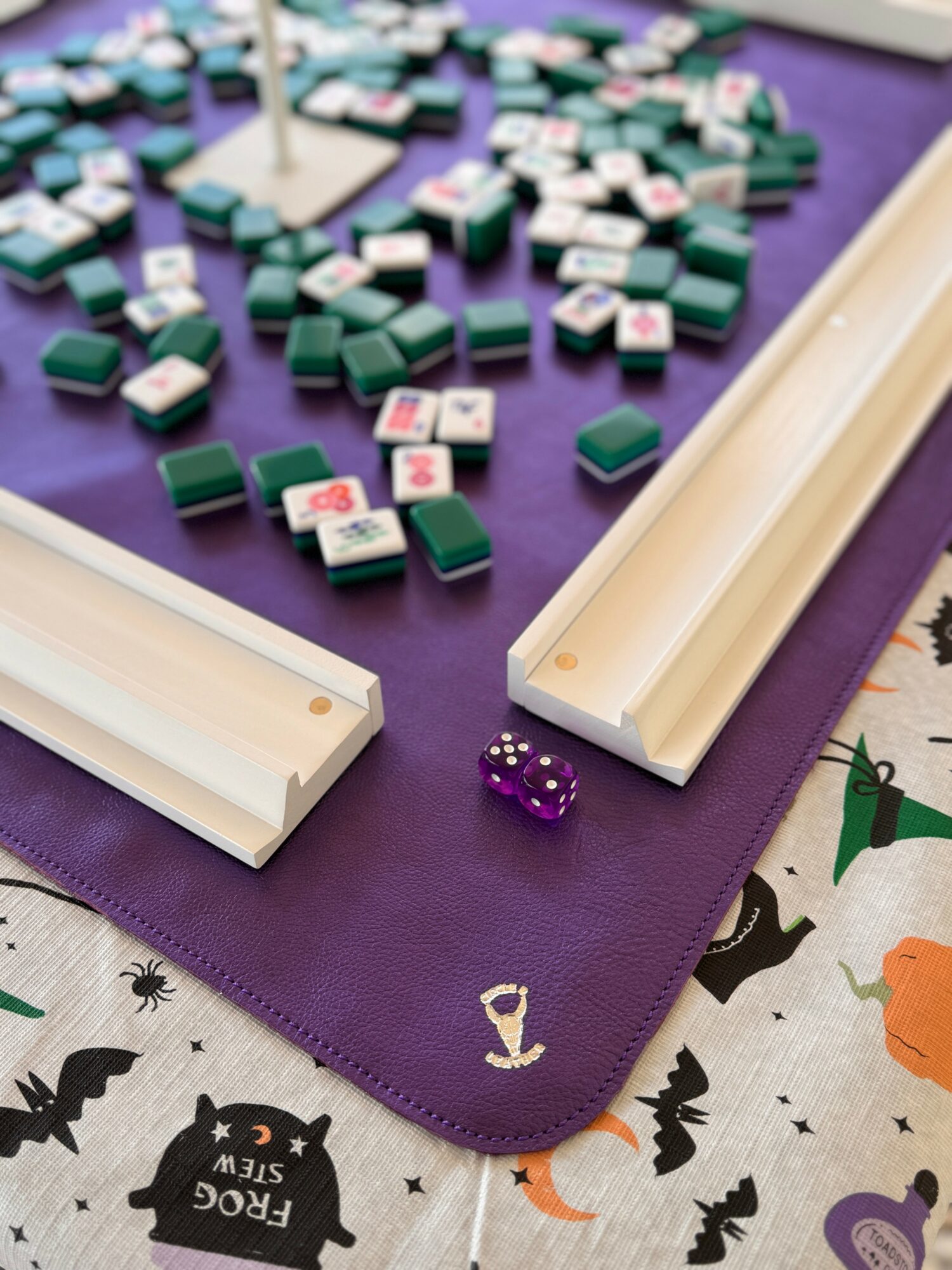 Mahjong game setup with tiles, purple dice, and a purple mat, on a cloth with bat and pumpkin patterns.