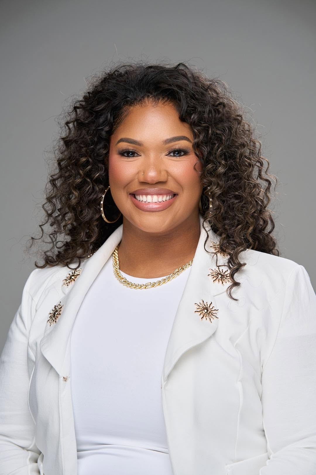 Portrait of a smiling woman with curly hair wearing a white blazer and gold jewelry, against a gray background.