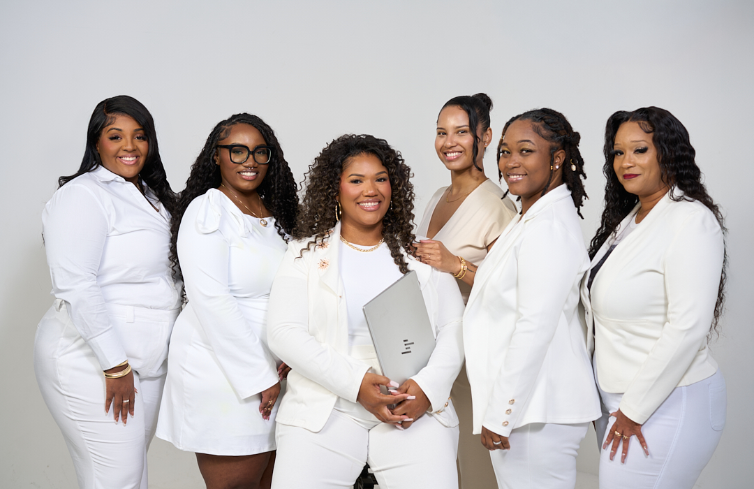 Six women in white outfits smiling, standing together against a plain background.