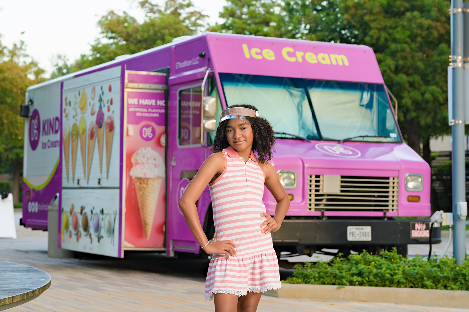 Young girl in pink and white striped dress standing in front of a pink ice cream truck outdoors.