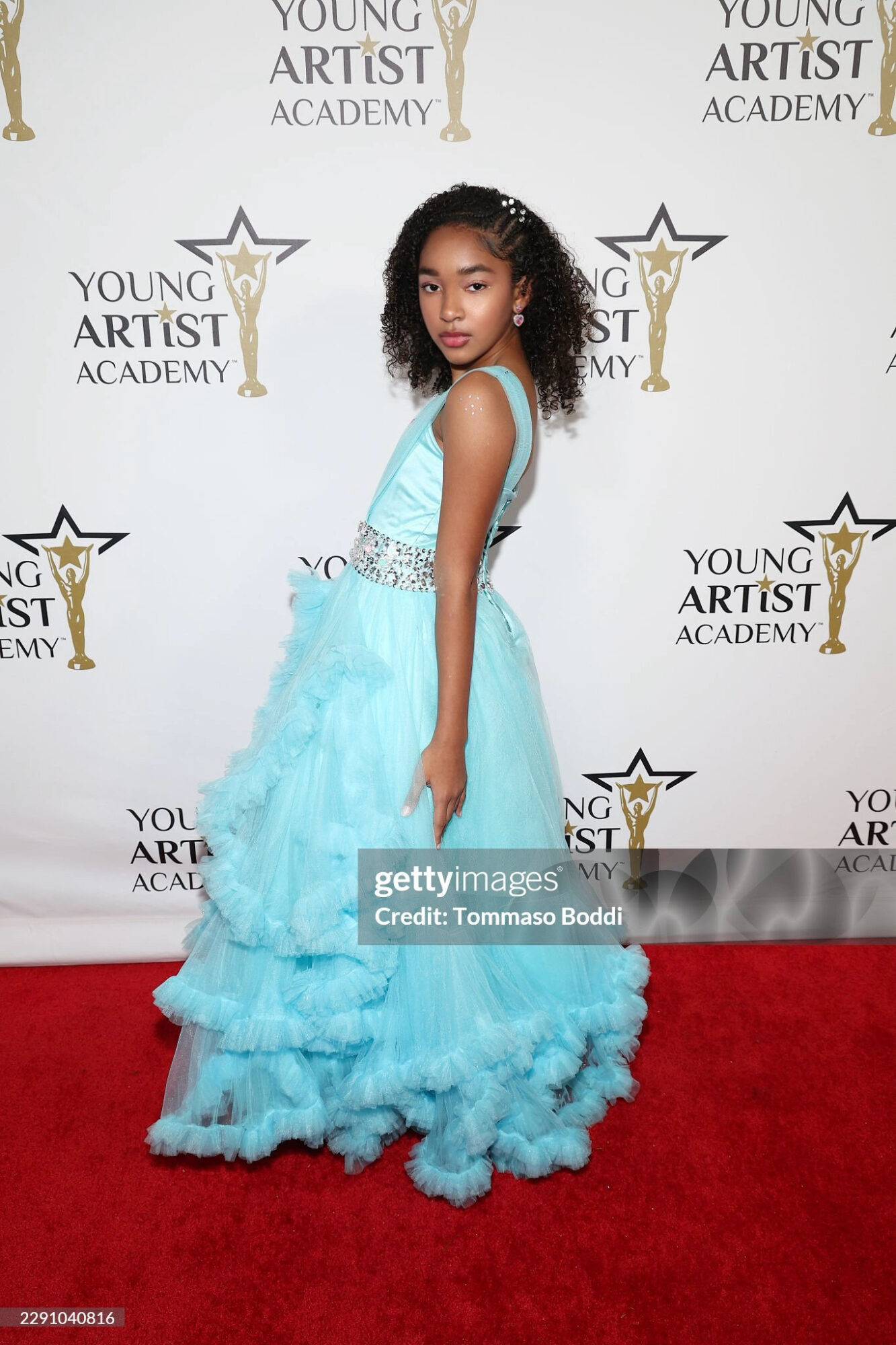 Young girl in a light blue gown with ruffles, standing on a red carpet at an event, with a backdrop featuring logos.