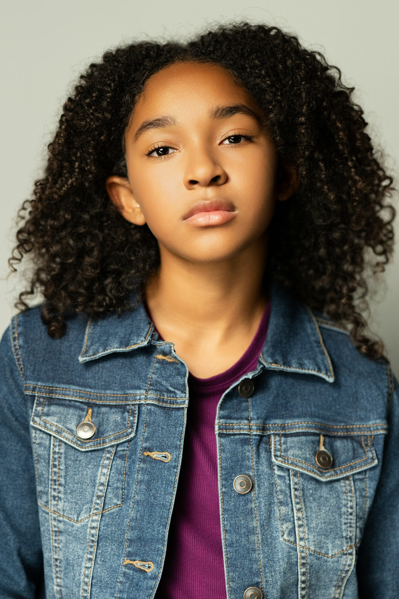 Young girl with curly hair wearing a denim jacket and purple shirt, looking at the camera.