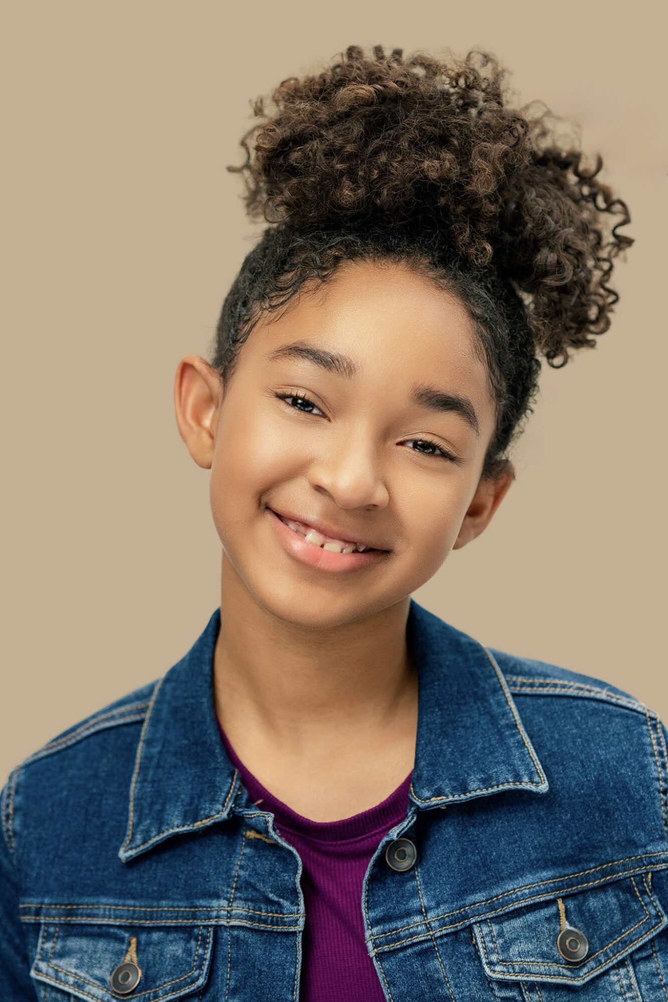Smiling girl with curly hair in a high ponytail, wearing a denim jacket and purple shirt, against beige background.
