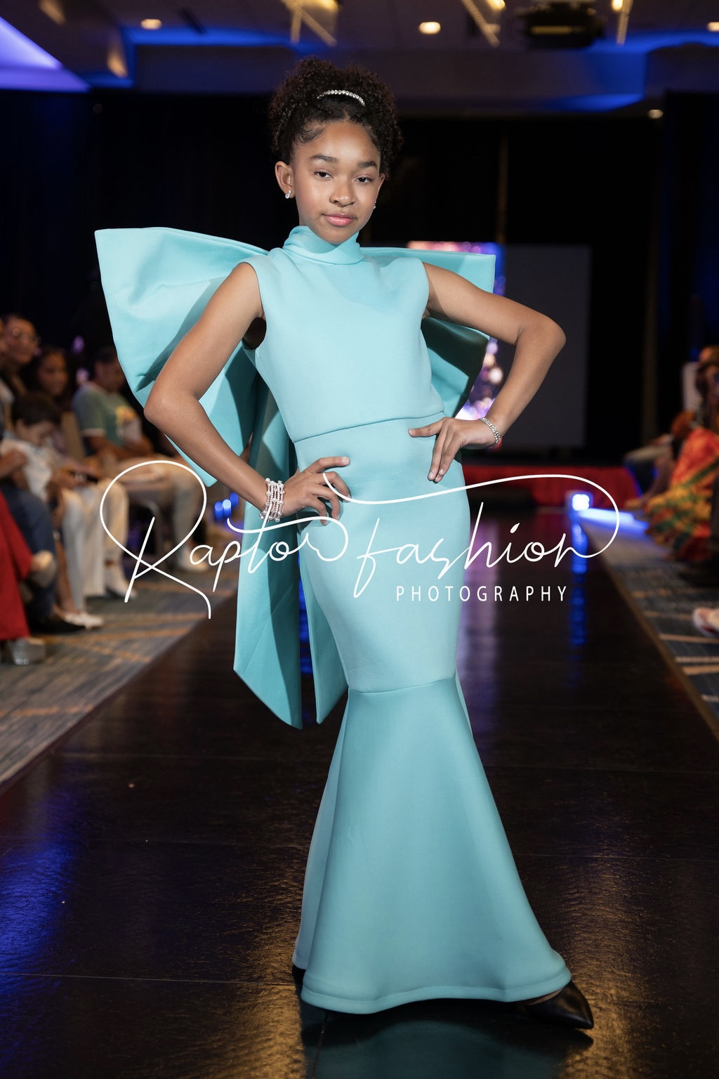 Young girl in a light blue gown with large bow, walking on runway with audience in background.