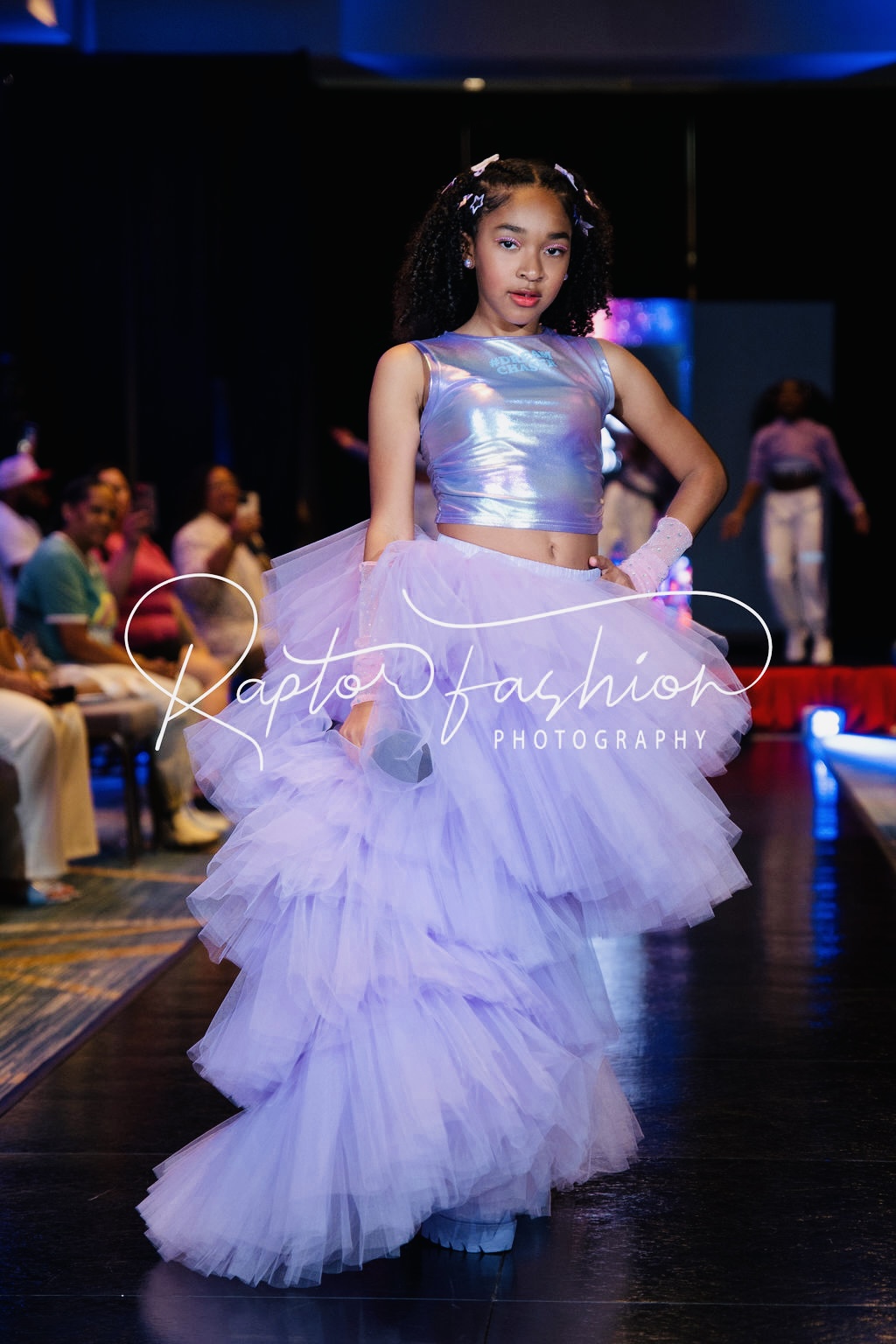 Young girl in a shiny purple top and layered lavender tulle skirt stands on a stage with an audience in the background.