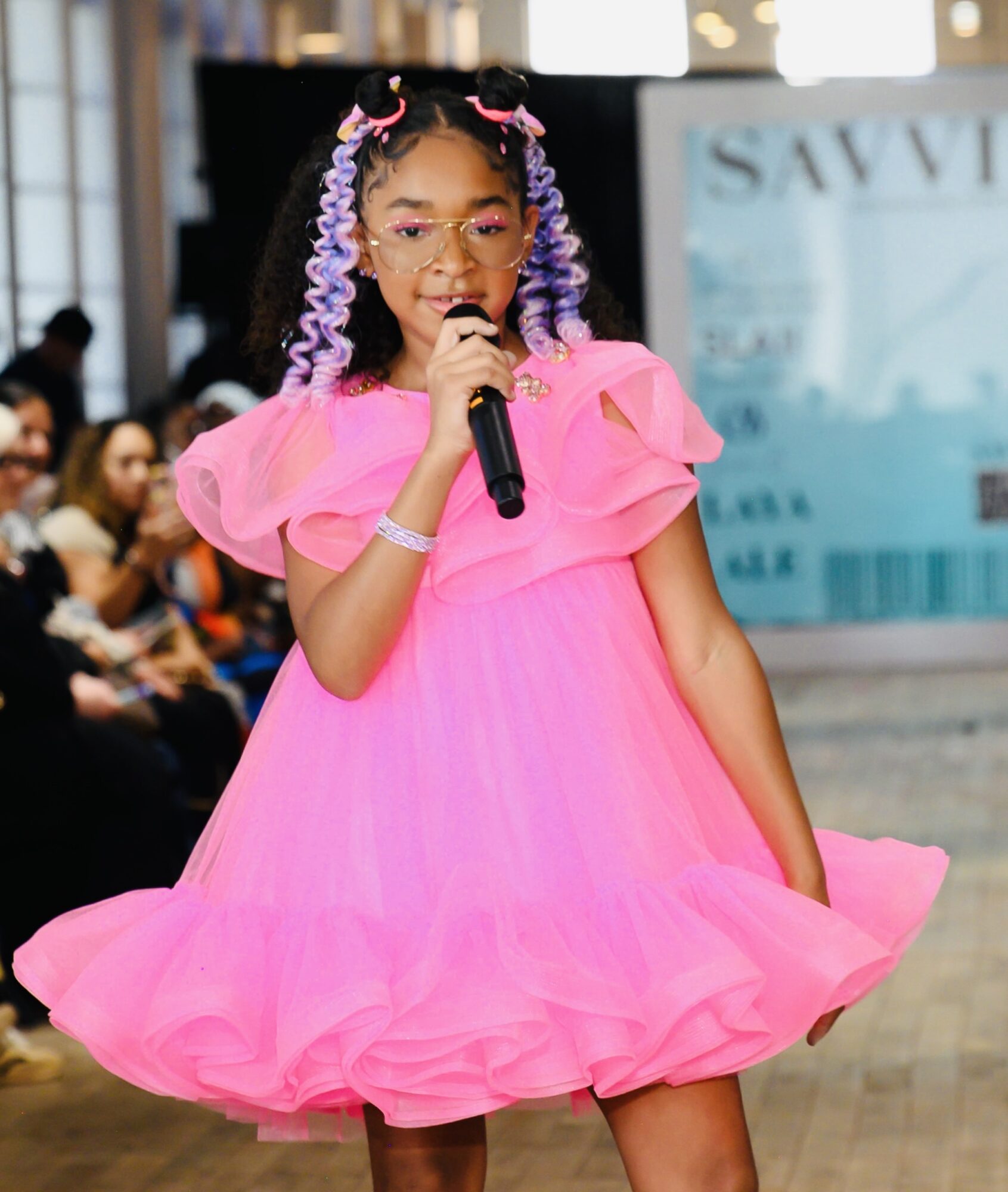 Young girl in pink dress holding microphone, performing on stage with audience in background.
