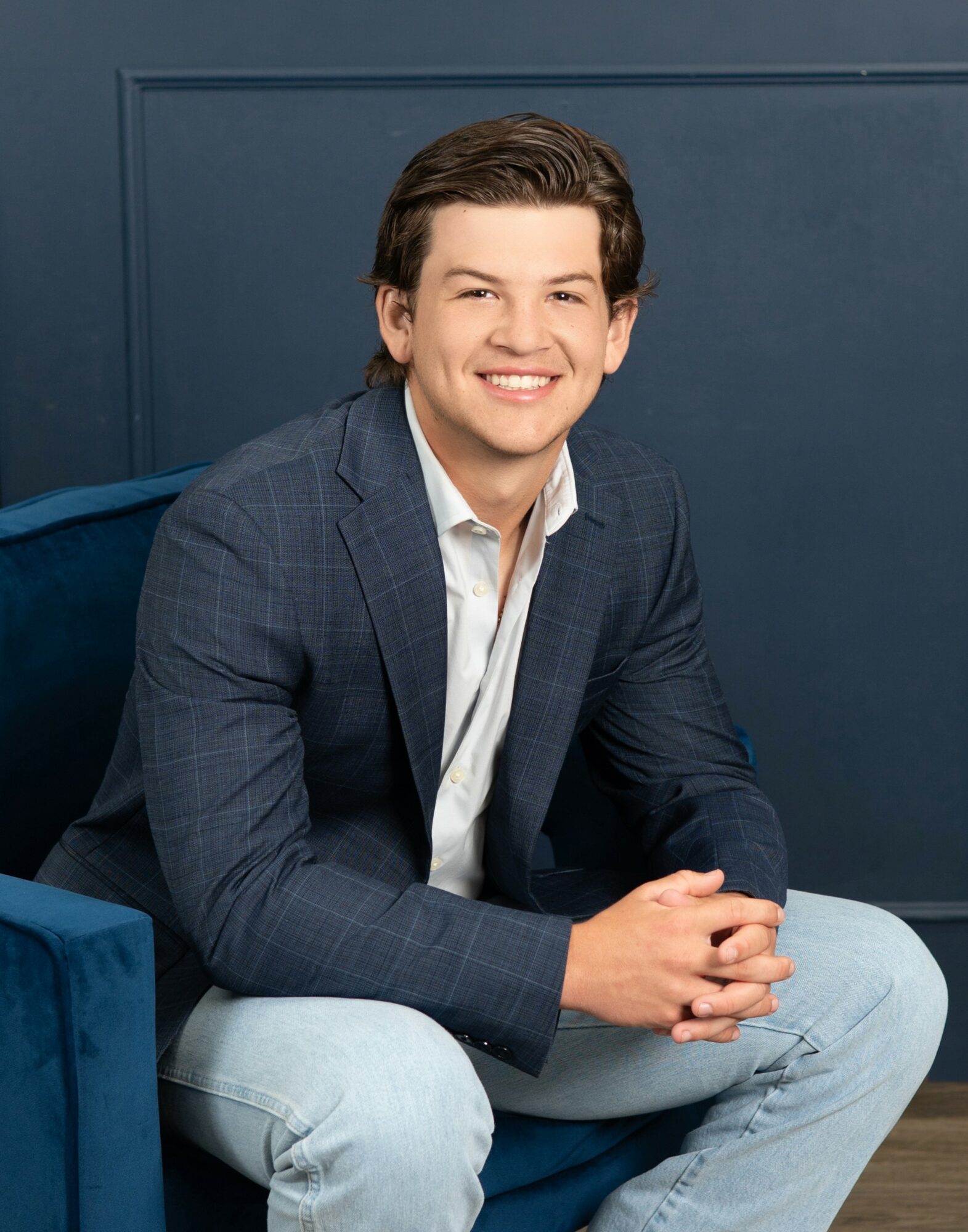 Man with dark hair smiling, sitting on a blue chair, wearing a dark blazer and light jeans, against a dark blue wall.