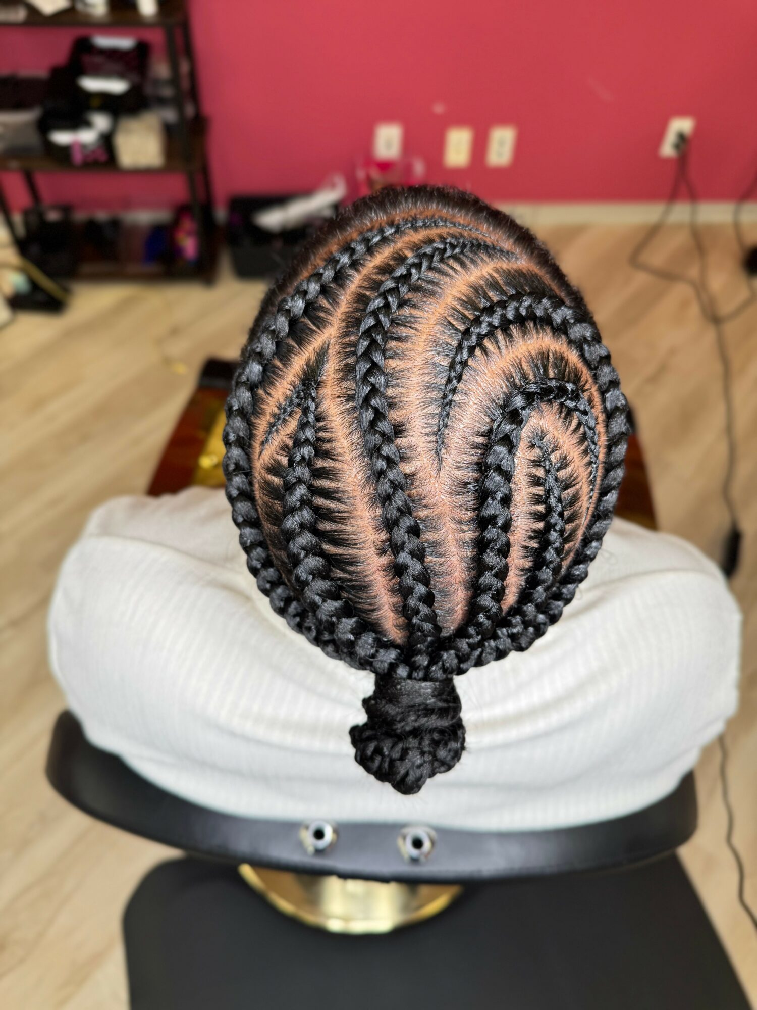Top view of a person's head with intricate braided hairstyle, sitting on a white cushion in a room with pink wall.