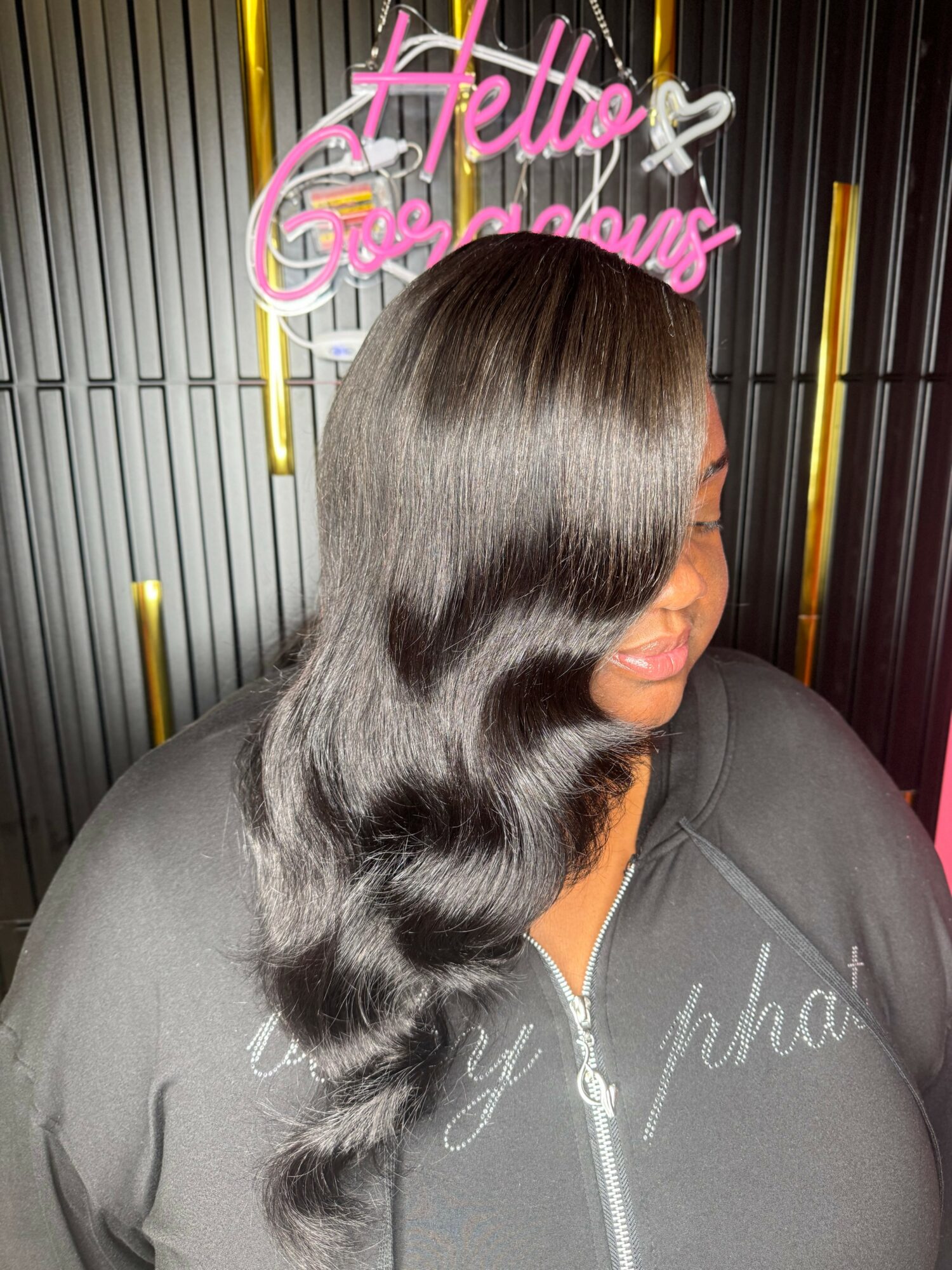 Side view of a woman with long, wavy, shiny black hair, standing in front of a neon sign. The sign reads 'Hello Gorgeous' in pink and white. She is wearing a dark zip-up hoodie. The background has vertical metallic stripes with yellow and pink accents.